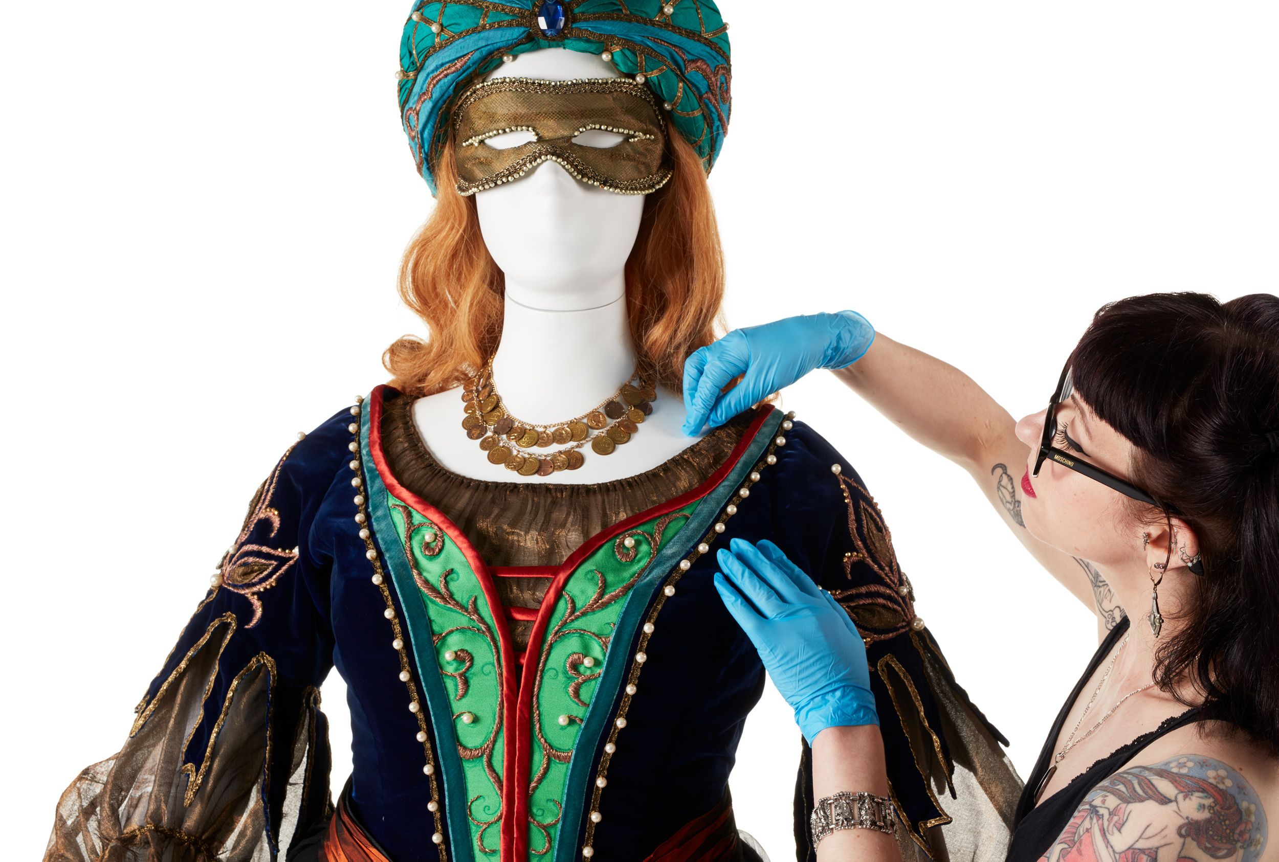 A conservator, wearing latex gloves, makes adjustments to the Lucrezia Borgia costume, and elaborate blue and green Renaissance-style costume, on a mannequin