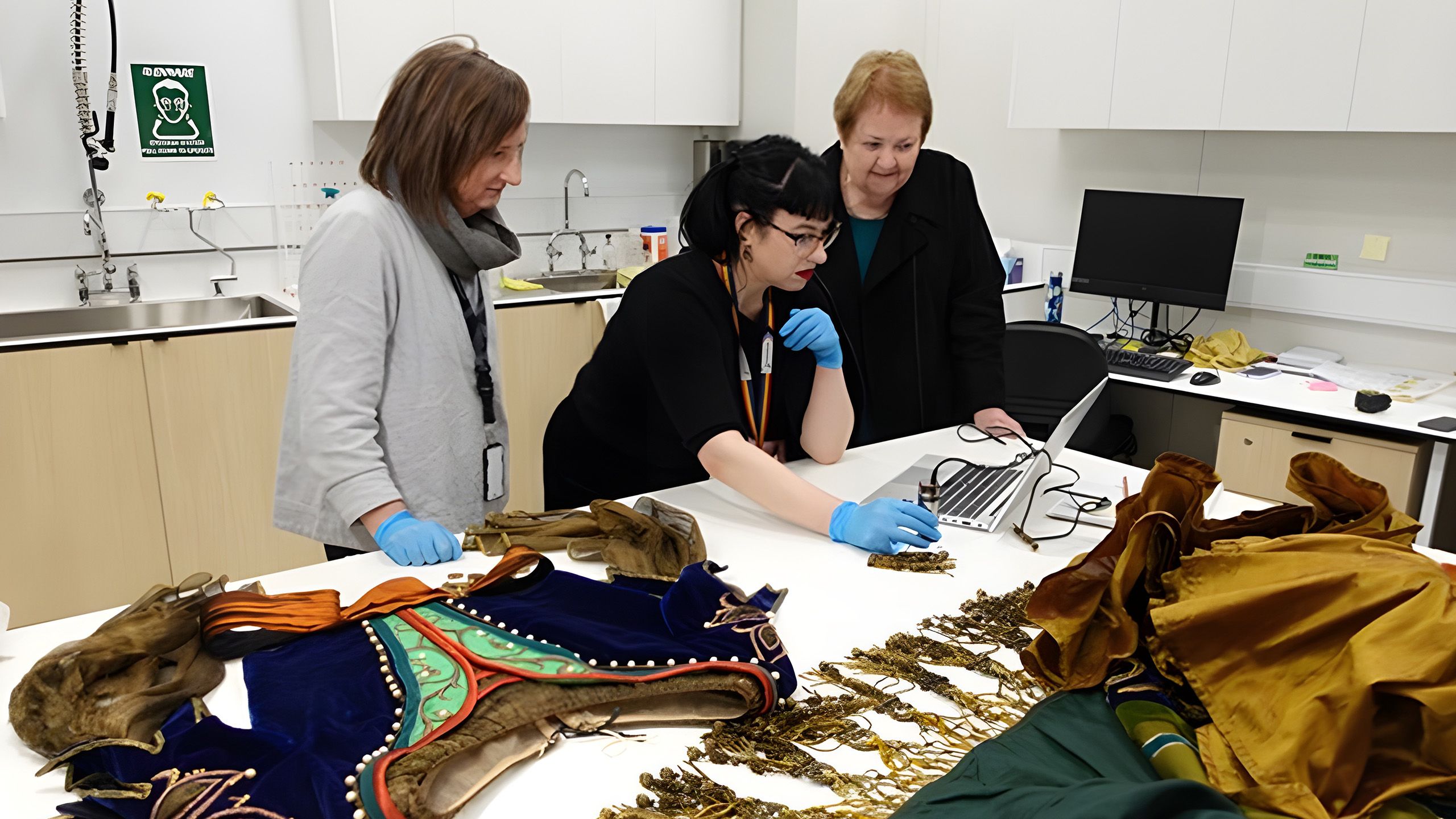 A conservator, conservation intern and donor, all looking at images on a laptop which has been placed on a table in a conservation lab which also has sections of the Lucrezia Borgia costume on it.