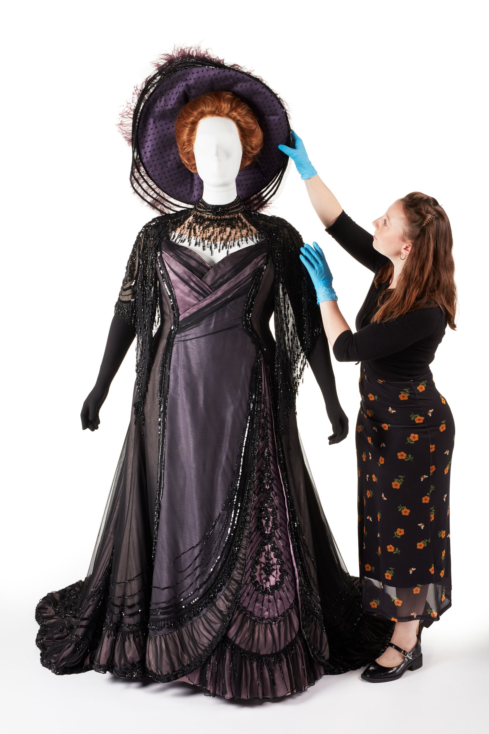 A conservator, wearing latex gloves, makes adjustments to the Merry Widow costume, a richly detailed purple dress and matching hat, on a mannequin