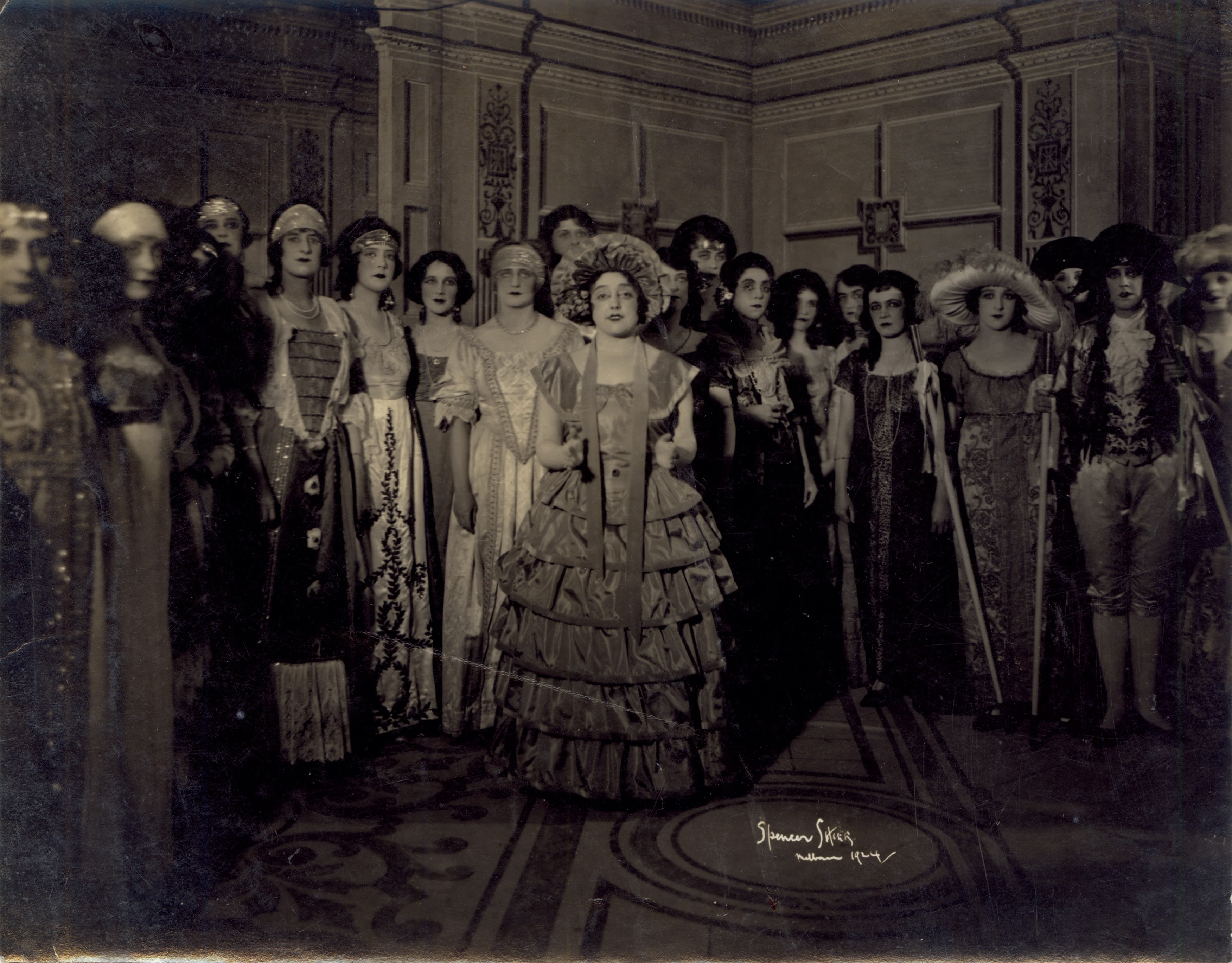 Photograph of 20 female performers on stage. They are all wearing ornate embroidered costumes with headdresses and headbands, looking directly at the camera. Toti Dal Monte is in the middle. She is wearing a floor-length tiered ruffle dress with a peter-pan neckline and large bonnet hat. Her hand are up at around her waist with her palms open. She is half smiling at the camera. 
