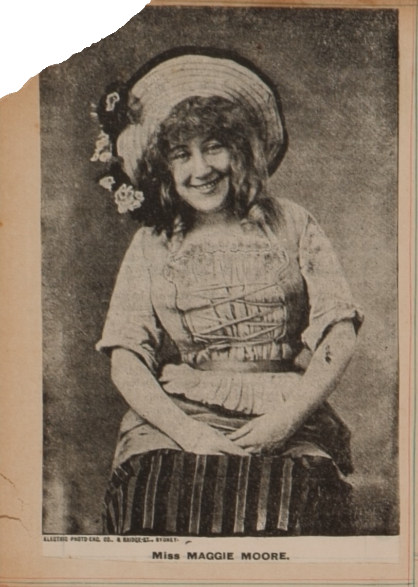 Photograph of woman sitting. She is wearing a dress with a rope corset at the front, and sleeves rolled up to her elbows. She is wearing a straw hat with four black and white flowers on the right of the brim. Her hands are in her lap and she is smiling at the camera. Photograph is cut out from a newspaper and stuck into a scrapbook. 