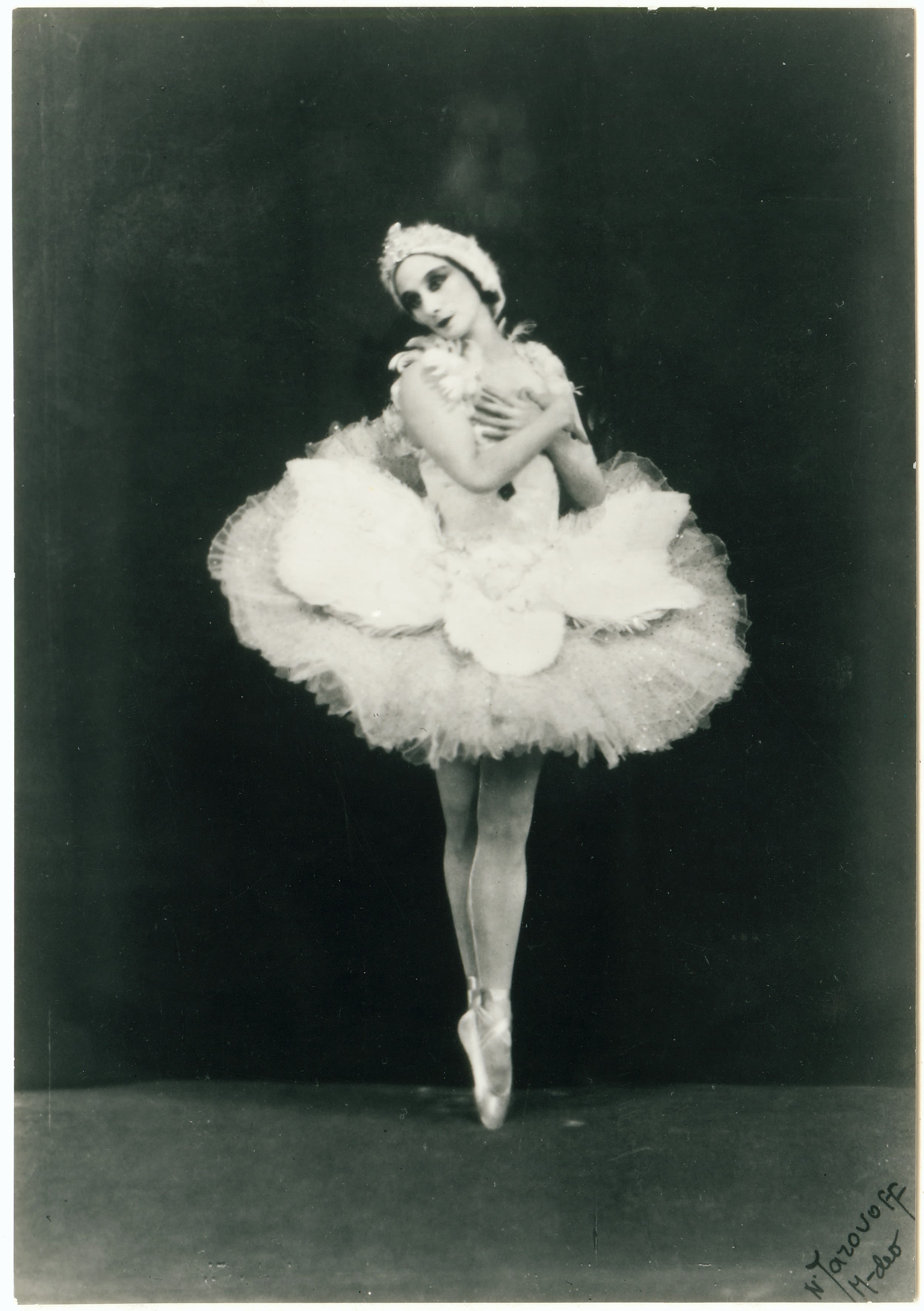 Full length portrait of a ballet dancer. She is wearing a white tutu made of tules and ruffles. She has her arms crossed on her chest and her feet are en pointe. She is wearing dark make up on her eyes and lips, and her head is tilted to the left. Her face is slightly angled to the left, and she is wearing a white headdress. She is in front of a black background. 