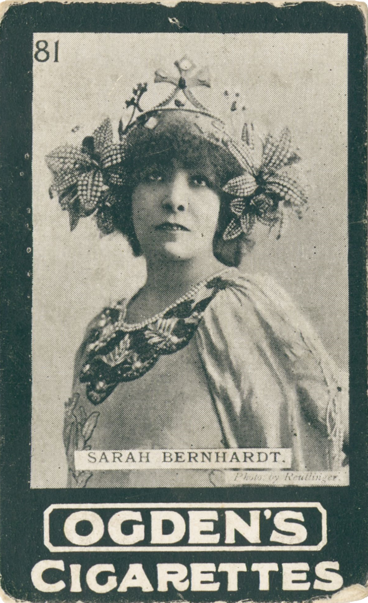 Cigarette card of a head and shoulders portrait of a woman. She is wearing a flowy white costume with an ornate embroidered neckline. Her headdress is made out of jewelled flowers on either side of her head, with a crown in the middle. She is faced directly towards the camera with her eyes looking up with a powerful facial expression. 'Ogden's Cigarettes' in block letters underneath the portrait.  