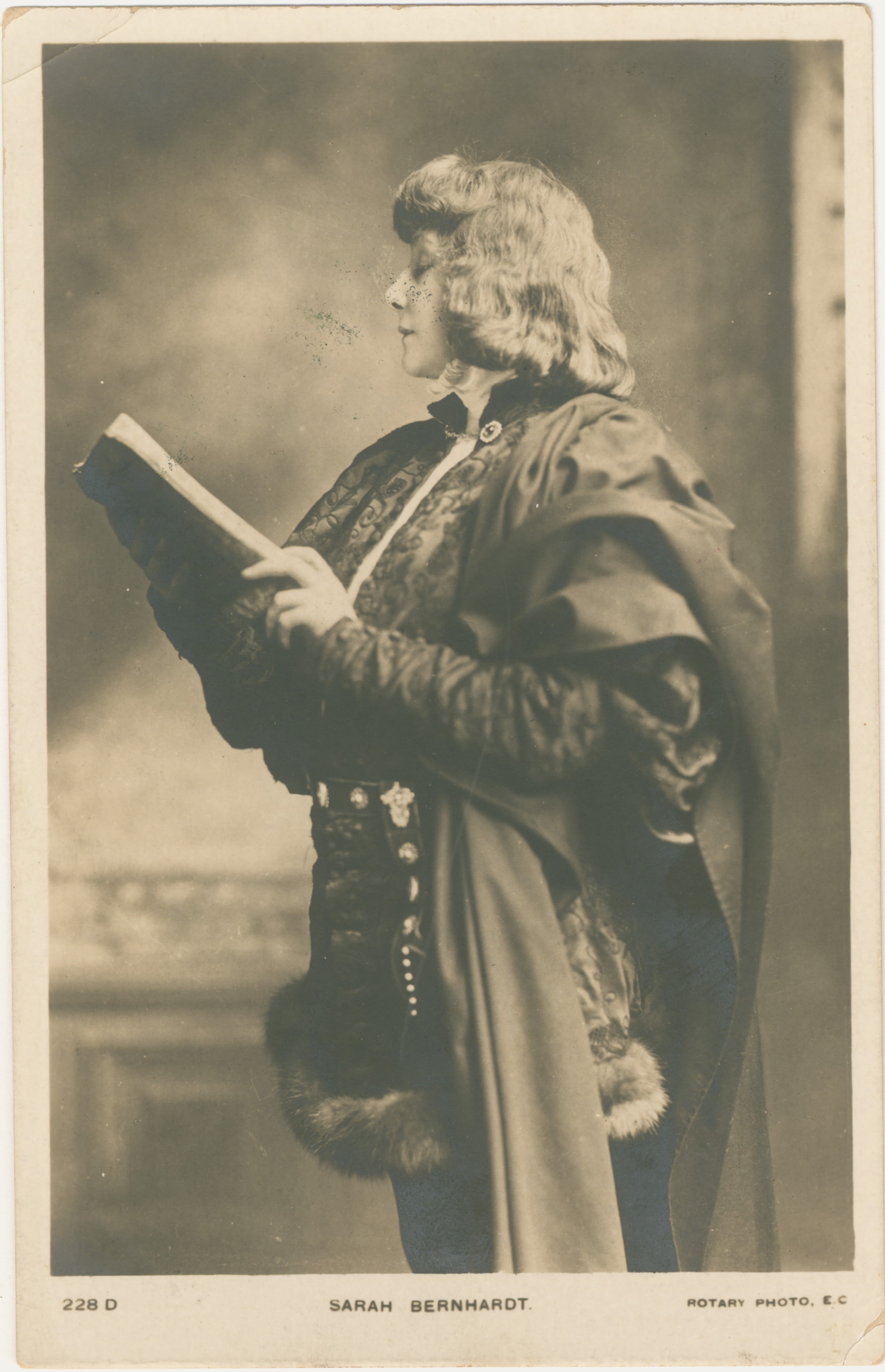 Portrait of a woman from her head to just above her knees. She is wearing a theatre costume which includes a hacket with fur trim on the bottom, a belt, and a shawl made from thick fabric sitting on her right shoulder. She is turned towards the left and is holding a book at chest-height. She is looking at the pages of the book. 