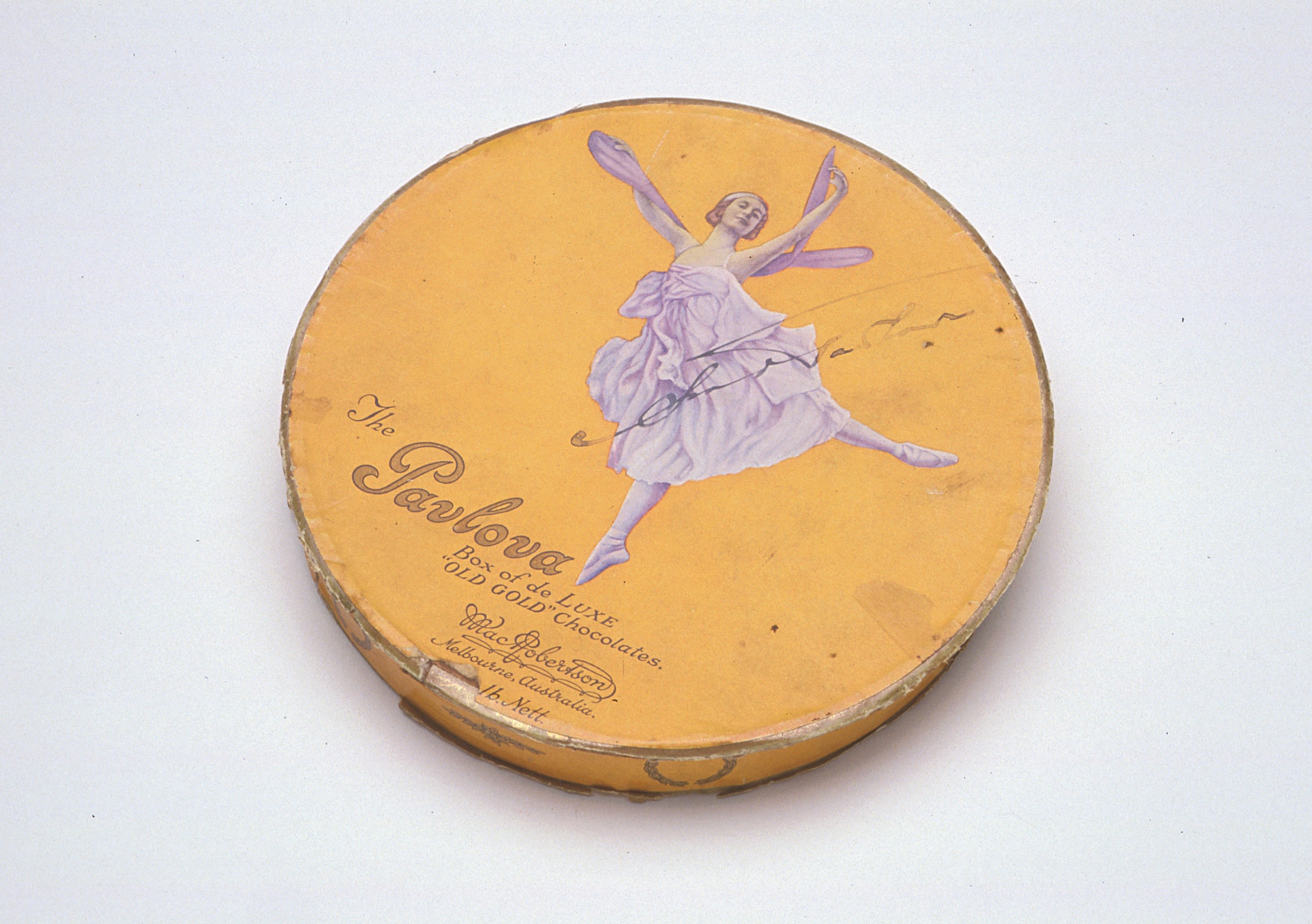 Photograph of a round metal chocolate box from the early twentieth century. The chocolate box is yellow with an image of a ballet dancer on the top. She is wearing a flowy knee-length dress and wings, and appears to be jumping in the air with her arms stretched up and out to the side. Her head is tilted to the right and she is half-smiling. There is an inscription on the bottom left: "The Pavlova Box of de LUX "Old Gold" Chocolates. MacRobertson. Melbourne, Australia. 1b.Nett. Signed by Anna Pavlova across the image of the ballet dancer. 