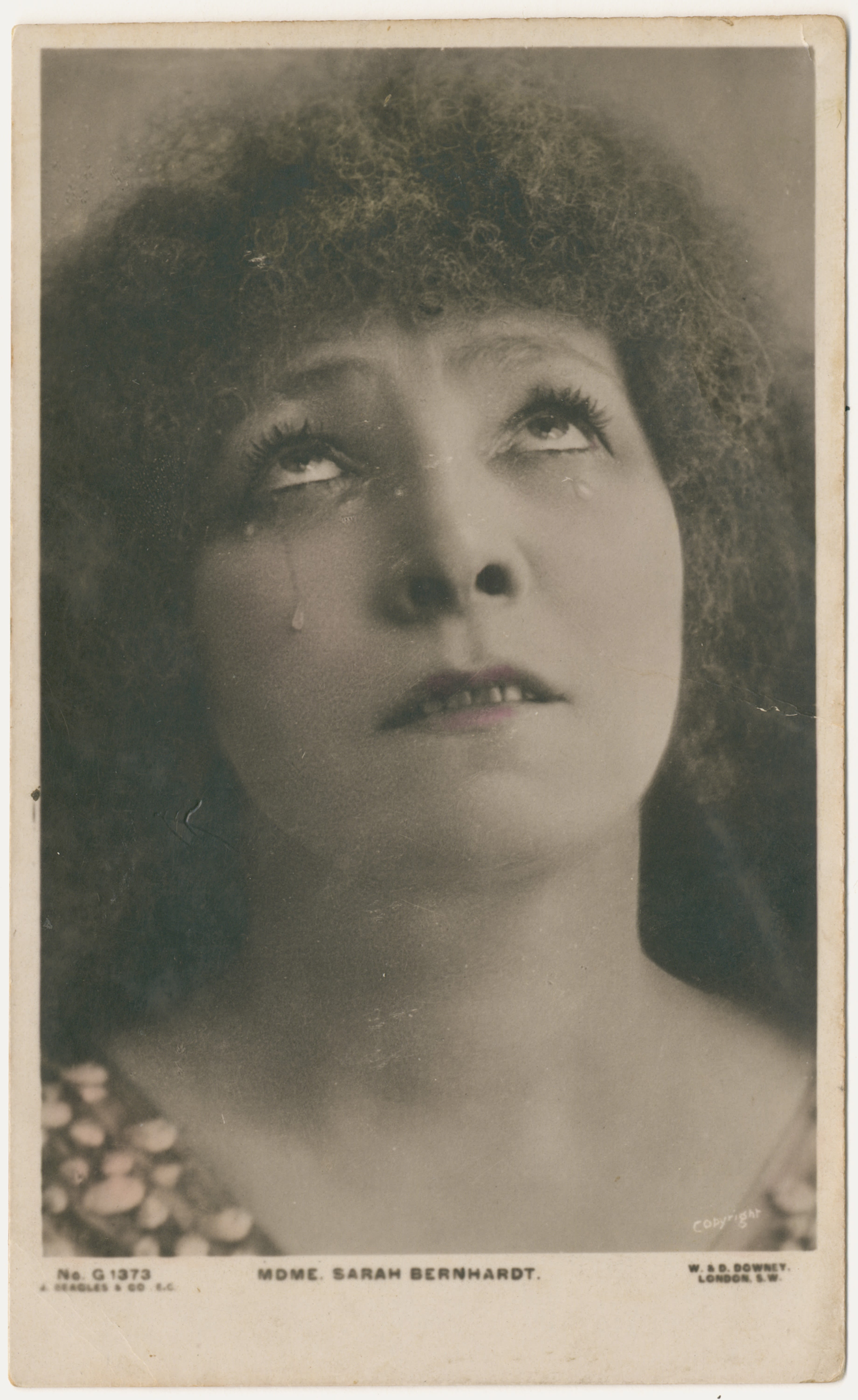 Close up head and neck portrait of woman. She is wearing a garment with a beaded neckline, and is face-on looking up towards the sky with a somber expression. Dramatic tears fall down her left cheek and past her eyelids. She has curly hair which covers her forehead. 