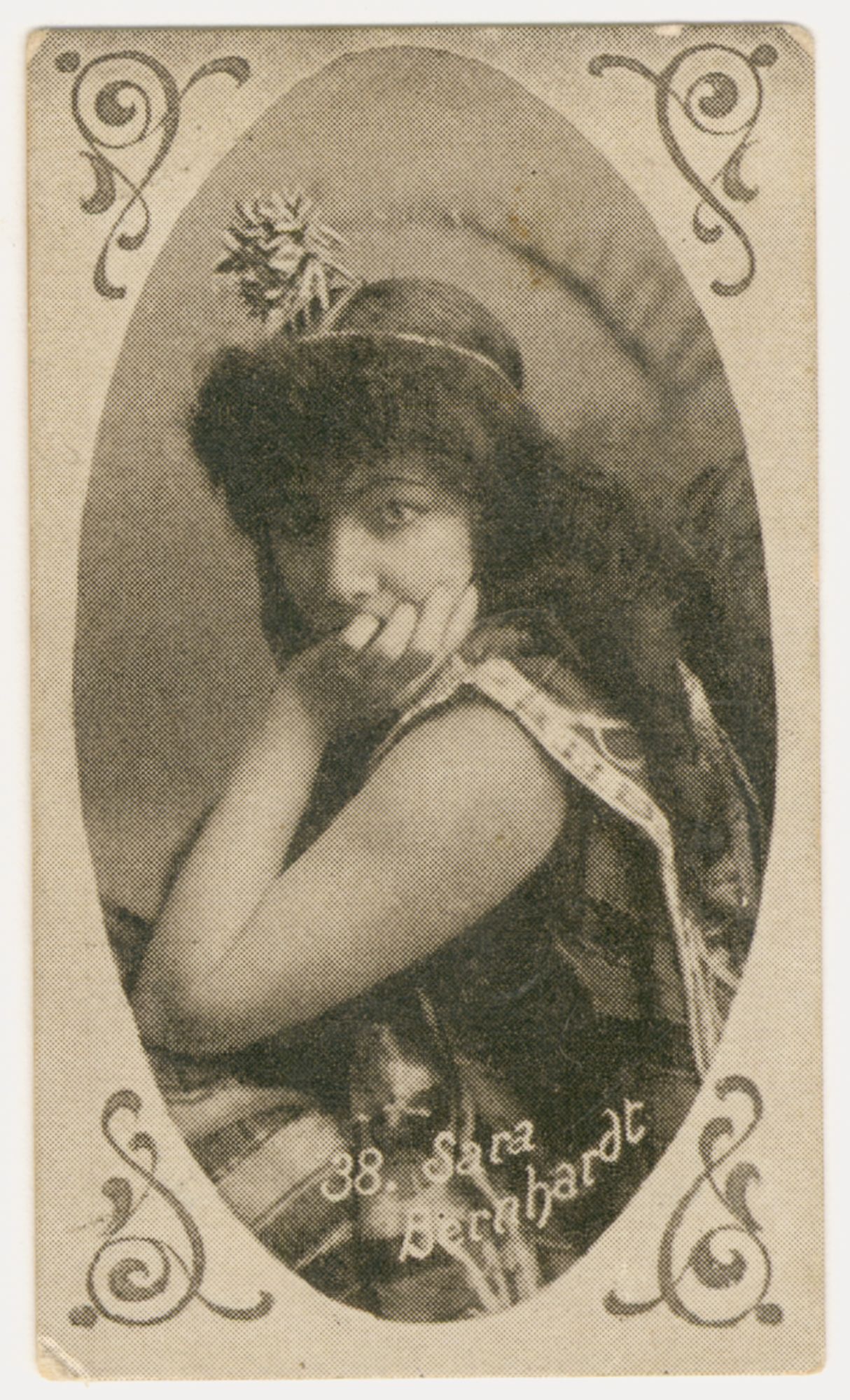 Portrait of a woman. She is crouched down, only her head, and shoulders are in frame. Her body is turned to the left, with her head facing the camera and her right arm folded with her right hand on her chin covering her mouth. She is wearing a striped theatre costume with a small headdress. She is looking direcrtly at the camera. Photo is in an oval frame, bordereed by swirly motifs on each corner. 