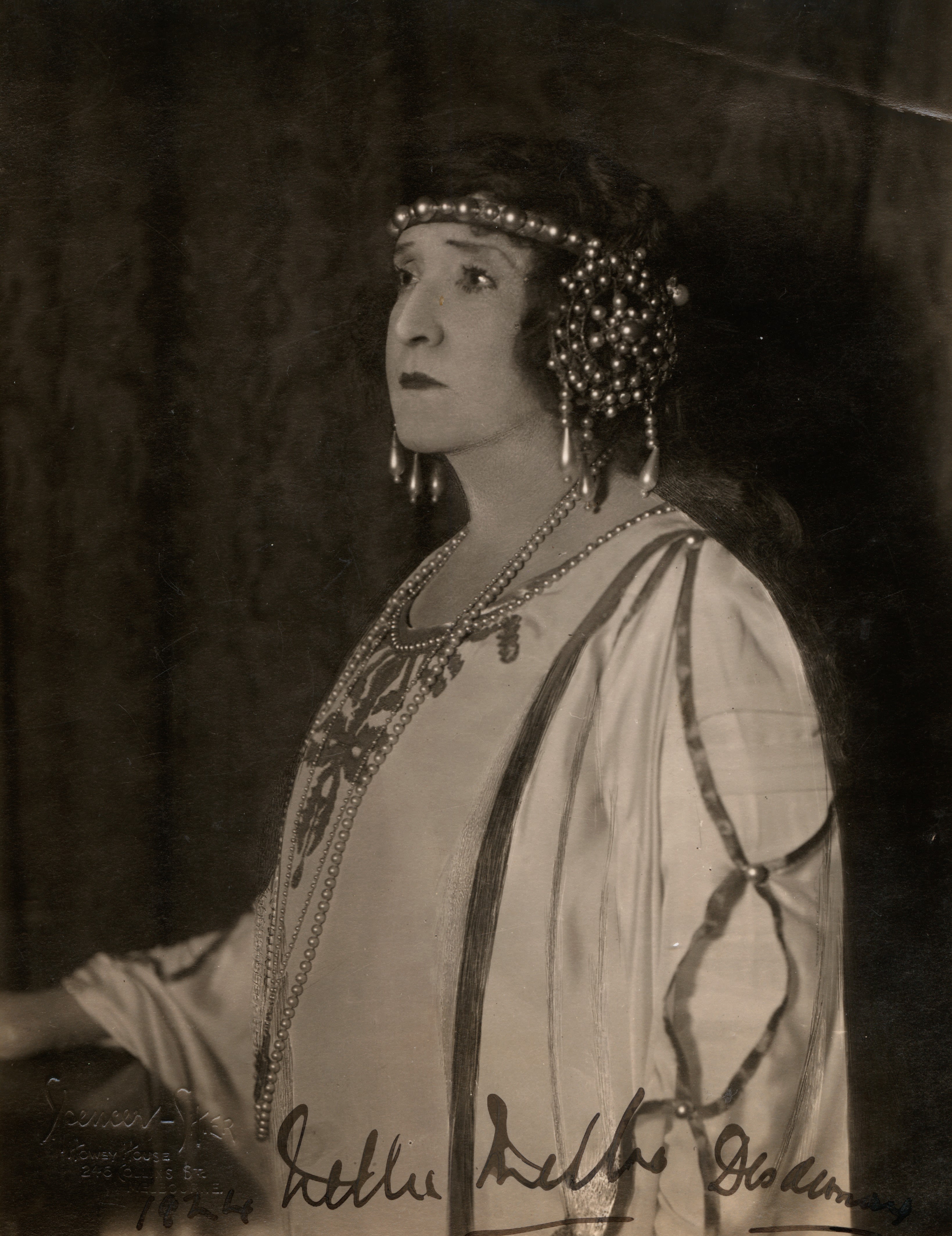 Head and upper-body portrait if a woman. She is wearing a headdress made of pearls, and a cream flowy costume with dark fabric strip accents and pearls on the sleeves. The costume has an ornately detailed neckline. She is wearing a long pearl necklace. She is turned to the left and looking out into the distance with a pensive look on her face. Signed 'Nellie Melba Desdemona' at bottom-right of the image in black cursive handwriting. 