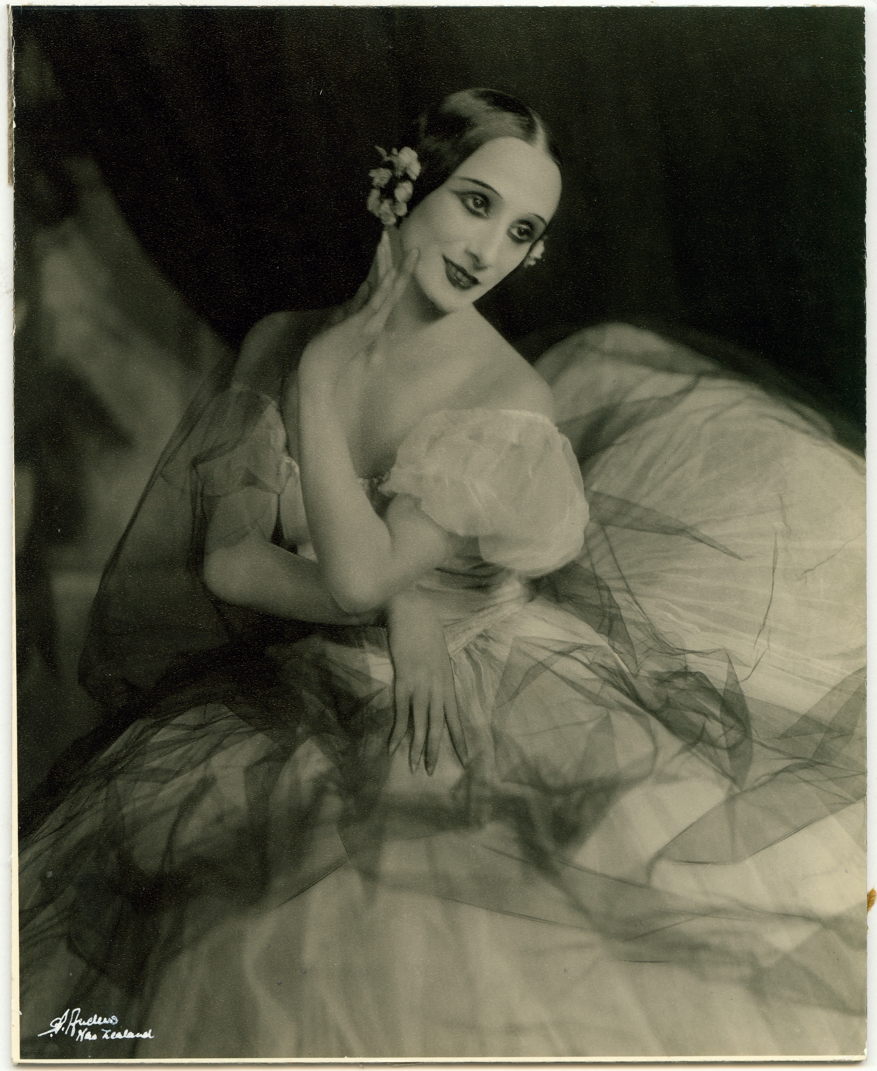 Head and shoulders portrait of a woman with a large tutu-like skirt filling up most of the frame. She is twisted to the left with her arms folded and her right hand raised up next the left side of her face. The white dress is off the shoulder with puffy sleeves, covered in black sheer tulle. Her hair is slicked into a middle-part with two small round flower headpieces near both of her ears. She is looking to the right with her face in full view and is half-smiling. 