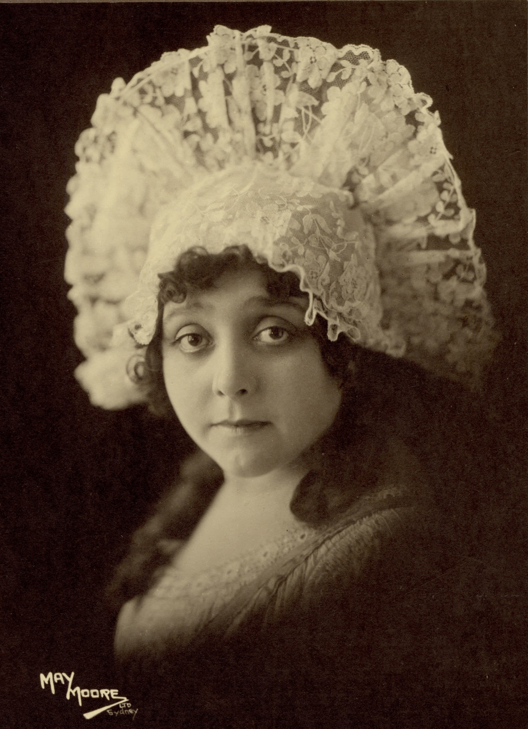 Head and shoulders portrait of a woman. She is wearing a tall headdress made of lace that fans out across to each of her ears. She is wearing a lace bodice. She has curly hair and is looking directly at the camera with a serious look on her face. 