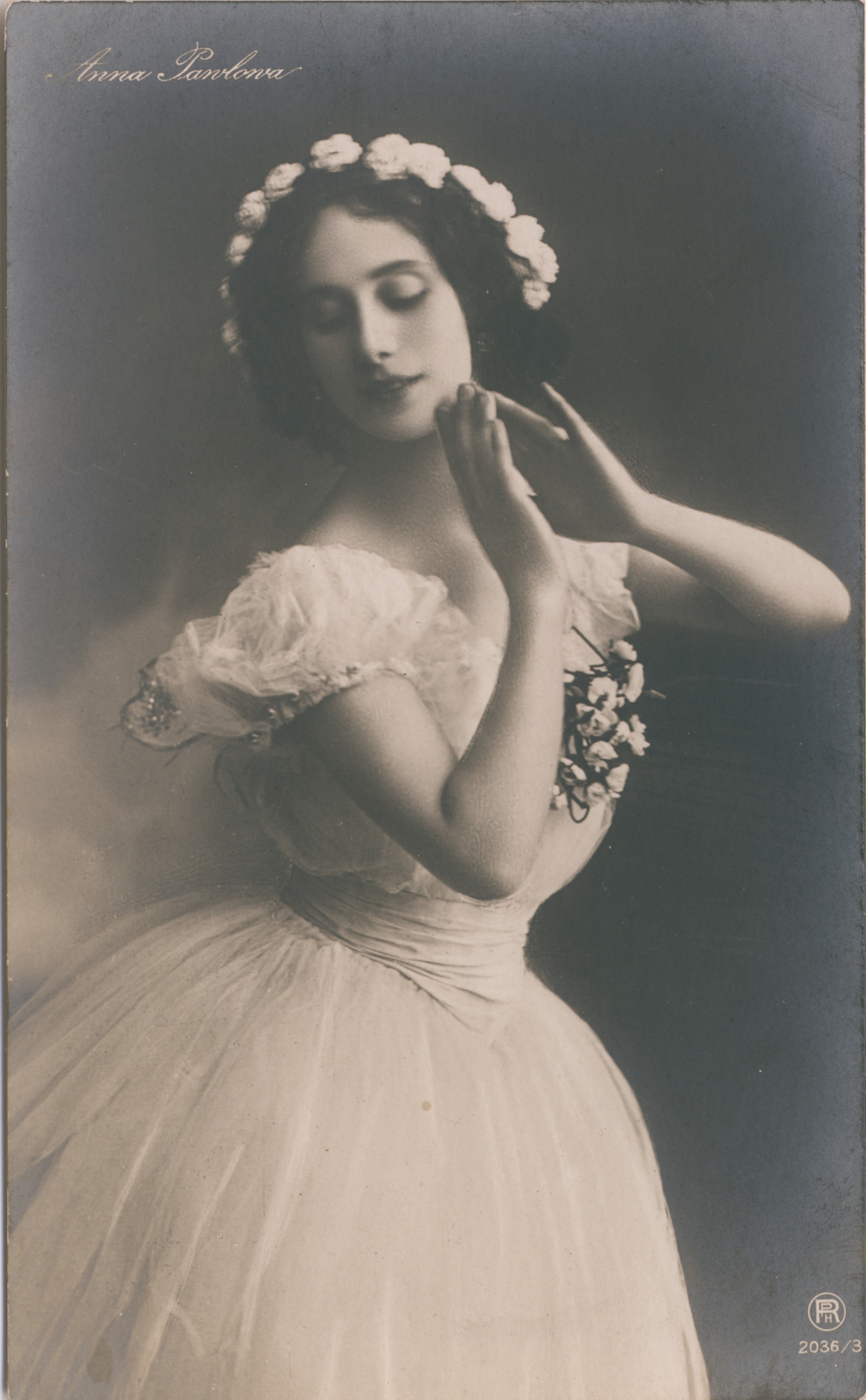 Portrait of a woman from her head to mid-thigh. She is wearing a ballet costume. The costume is a white dress with a tulle skirt and puffy sleeves, with a  flower-like decoration on her front chest. She is wearing a flower crown. Her arms are raised, elbows bent, with her hands just touching near her chin. She is looking towards the bottom left corner of the frame. 