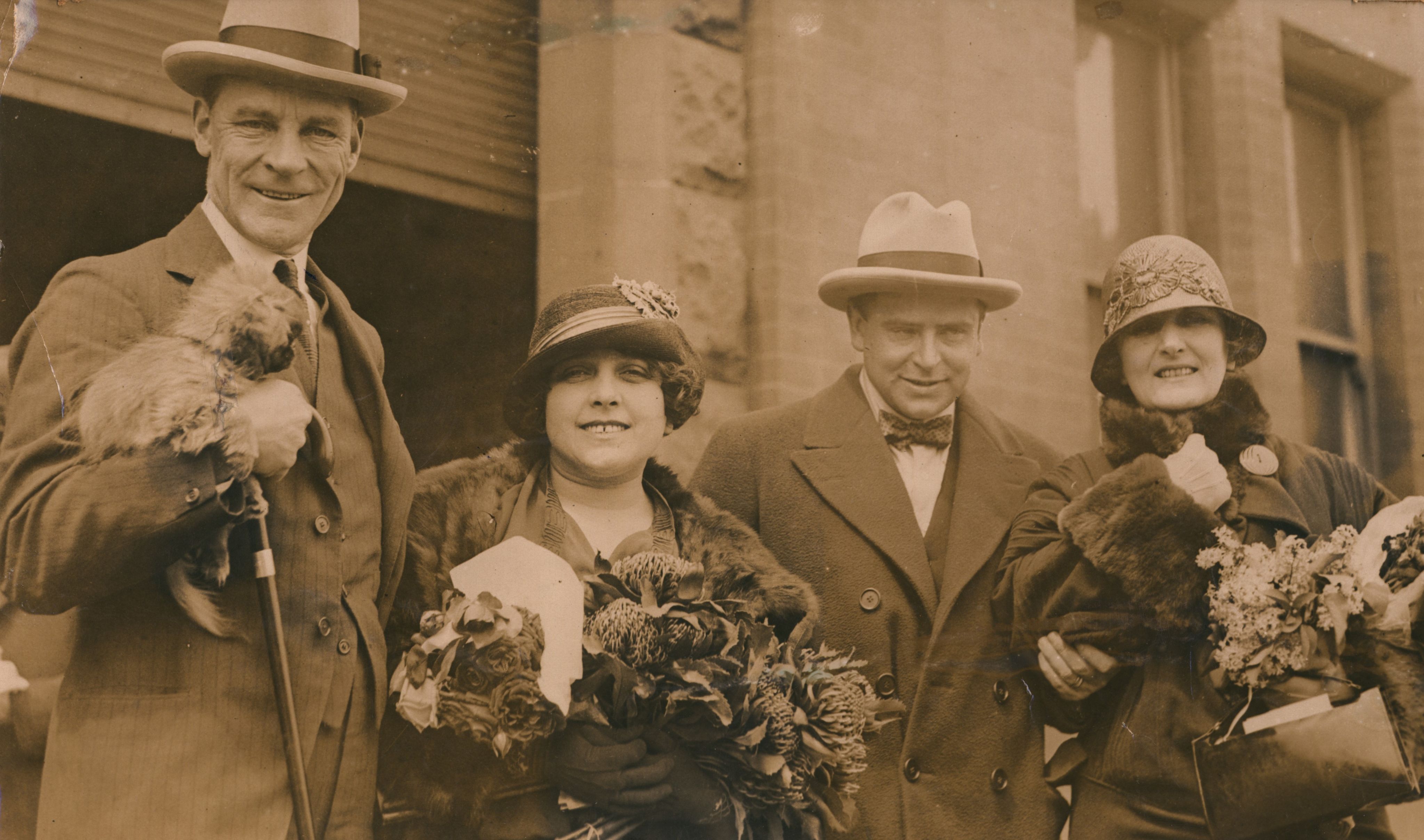 Sepia photograph of two men and two women. Man on the far left of the image is tall and wearing a three-piece suit and top hat, holding a walking stick and a small fluffy dog in his left arm. He is looking at the camera and smiling. Next to him is a short woman wearing a fur coat and holding three bouquets of flowers. She is wearing a hat with a ribbon tied in a bow across the hat. She is looking at the camer and smiling. Next to her is a man dressed in a suit and coat with a bow tie. He is wearing a top hat and smiling at the camera. His right hand is holding the elbow of the woman to his right. She is wearing a coat with fur trim on the cuffs and collar. She is wearing a bowler hat and holding boquets of flowers and a black rectangular handbag in her right hand. Her left hand is clutching the top of the fur collar. She is looking at the camera and smiling. All four people are standing behind a building with a garage door and windows. Photograph cuts off at around the figures' hips. 