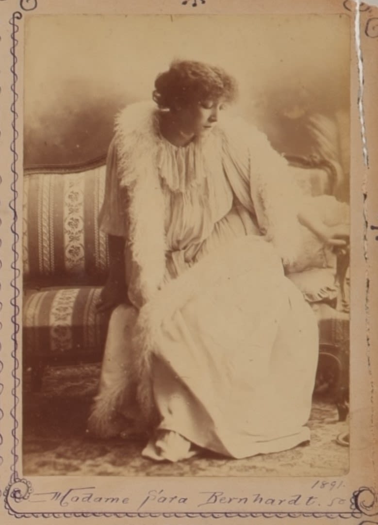 Full body portrait of a woman. She is sitting on a fabric striped couch wearing a flowy white theatre costume and feather boa. She is looking down towards the bottom right corner of the frame and her right hand is placed on the arm o the couch. Photograph is stuck onto scrapbook page. The photograph is decorated with a hand drawn lined border in blue pen. 'Madame Sarah Bernhardt' inscribed in blue pen at base of photograph. 