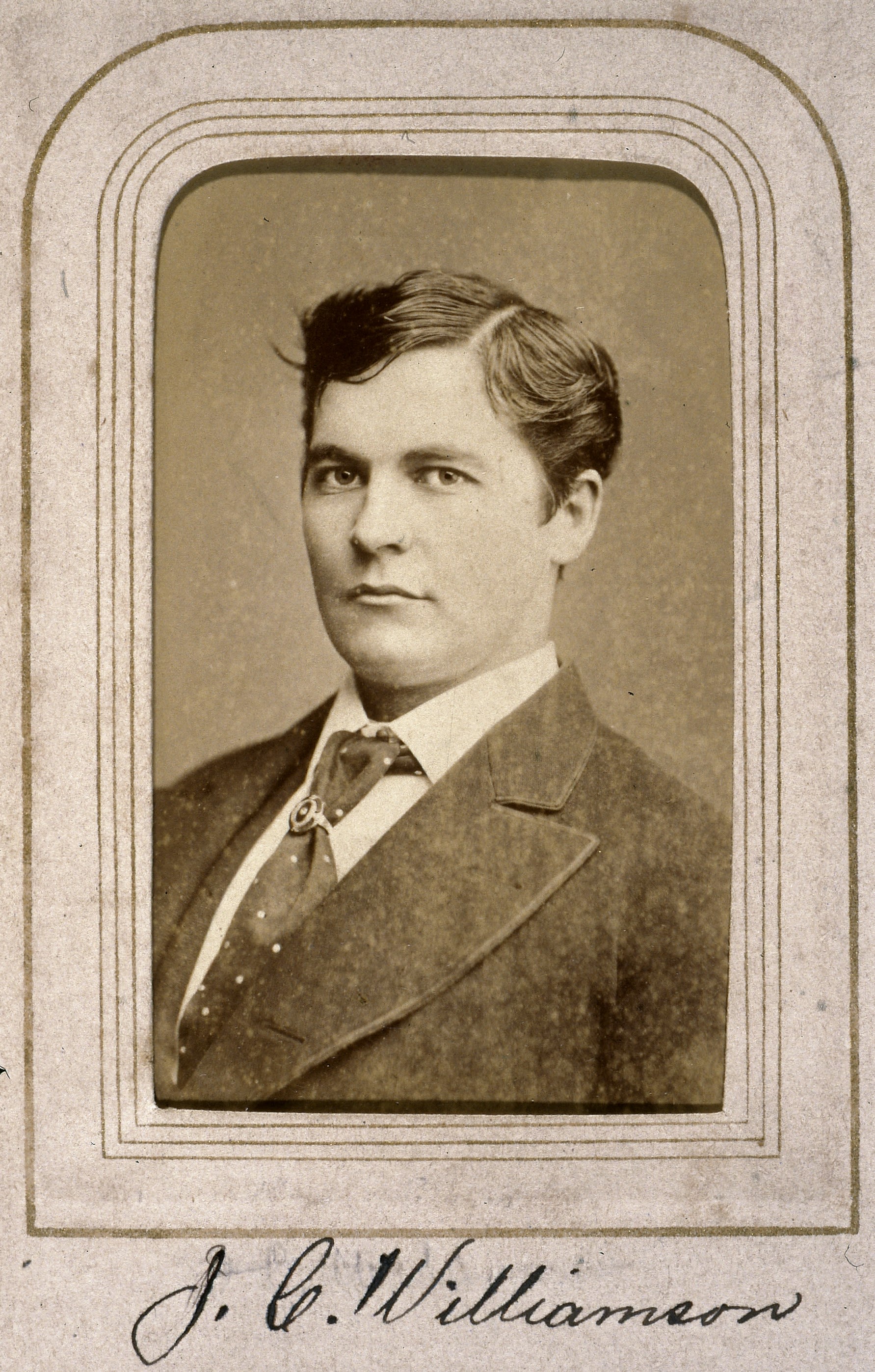 Head and shoulder portrait of a man wearing a suit looking at the camera. Photo stuck inside a scrapbook. Label under photo in cursive writing 'J.C. Williamson'. 