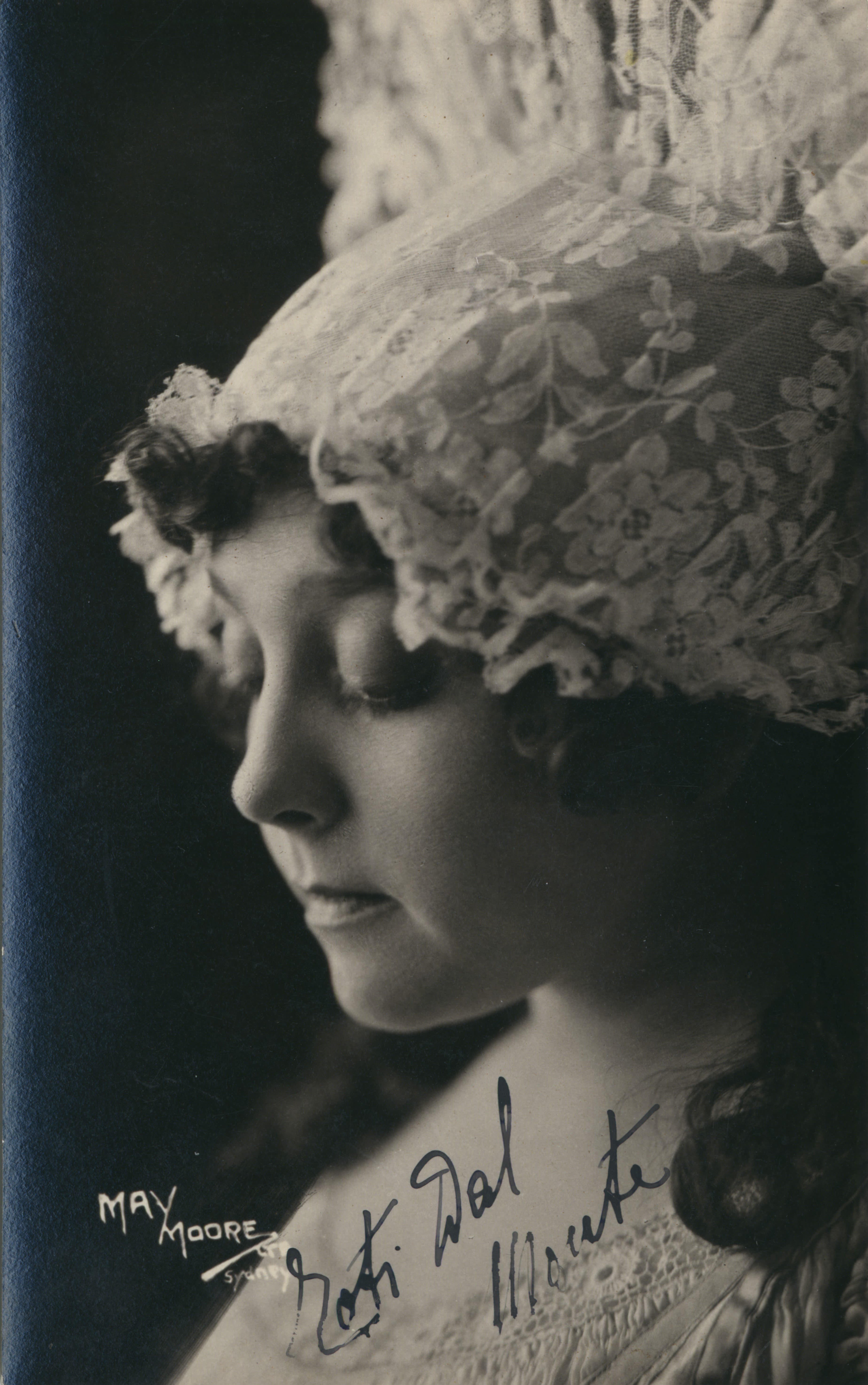 Head and shoulders portrait of a woman. She is wearing a tall headdress made of lace that fans out above her head. She is turned to the left and is looking down. 