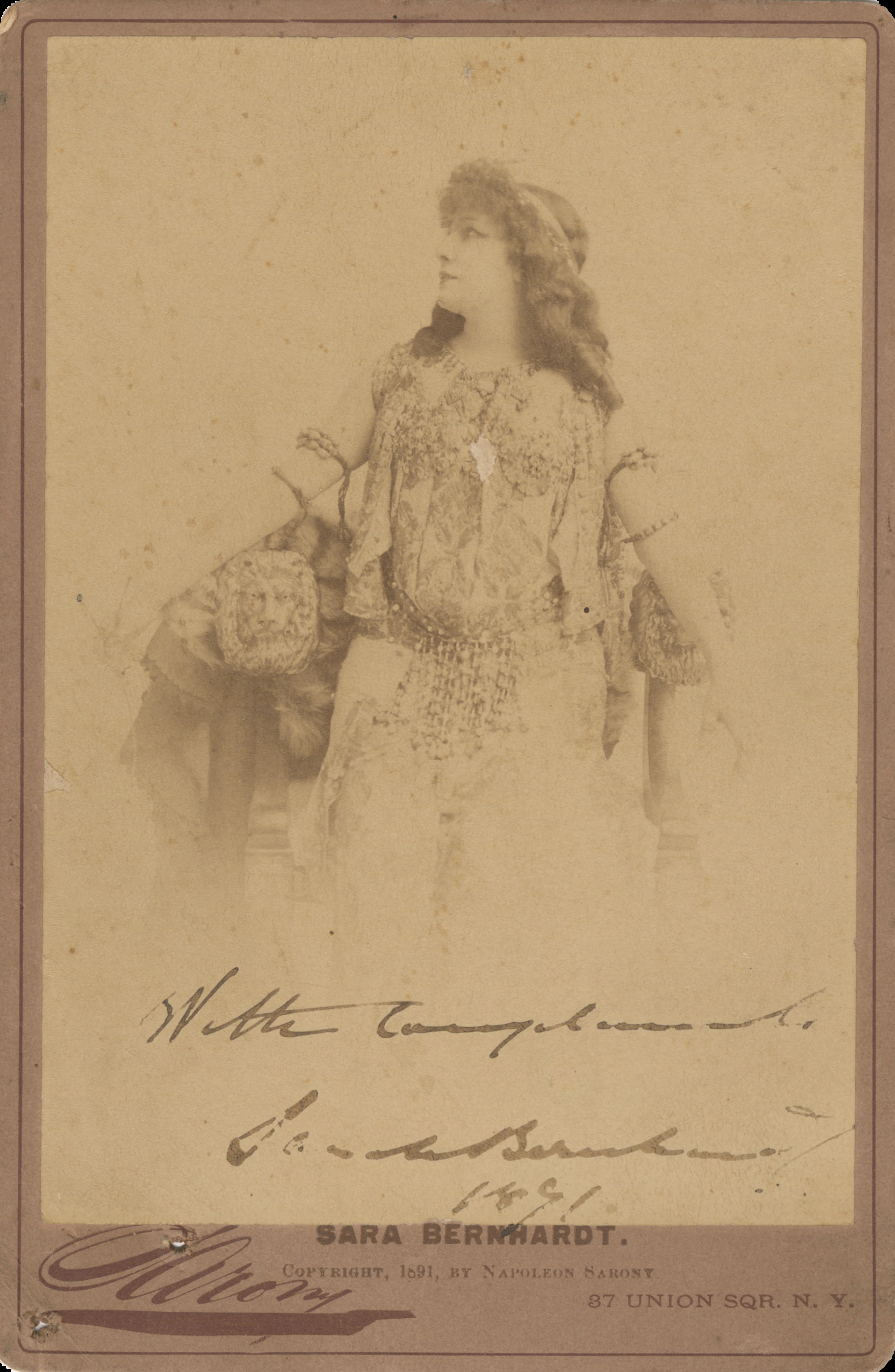 Faded portrait of woman in theatre costume. She is wearing a theatre costume heavily beaded in jewels, which include jewel cuffs on her upper arm. She is sitting on a large armchair with a statue of a lion on the left arm of the chair. She is looking across to her left showing her profile and long curly hair. Inscribed with a signature.
