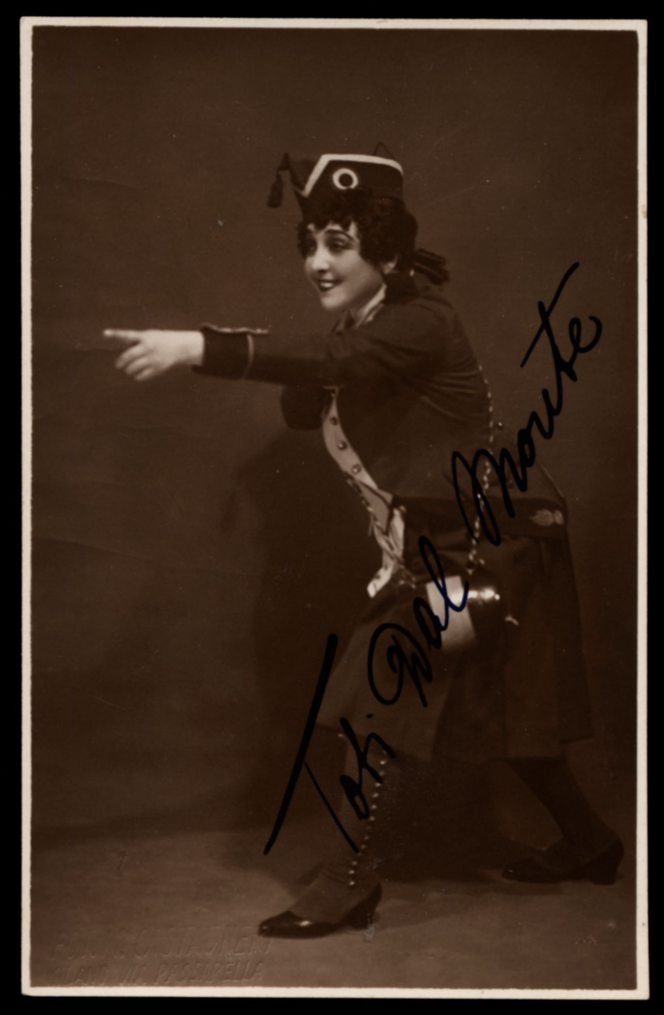 Full length portrait of a woman. She is dressed in a military style costume with a sailor hat. Her right leg is across her body to the left, and she is looking towards the left and smiling. Her right arm is pointed across her body to the left. Handwritten cursive signature in black pen is written diagonally up over her body. 