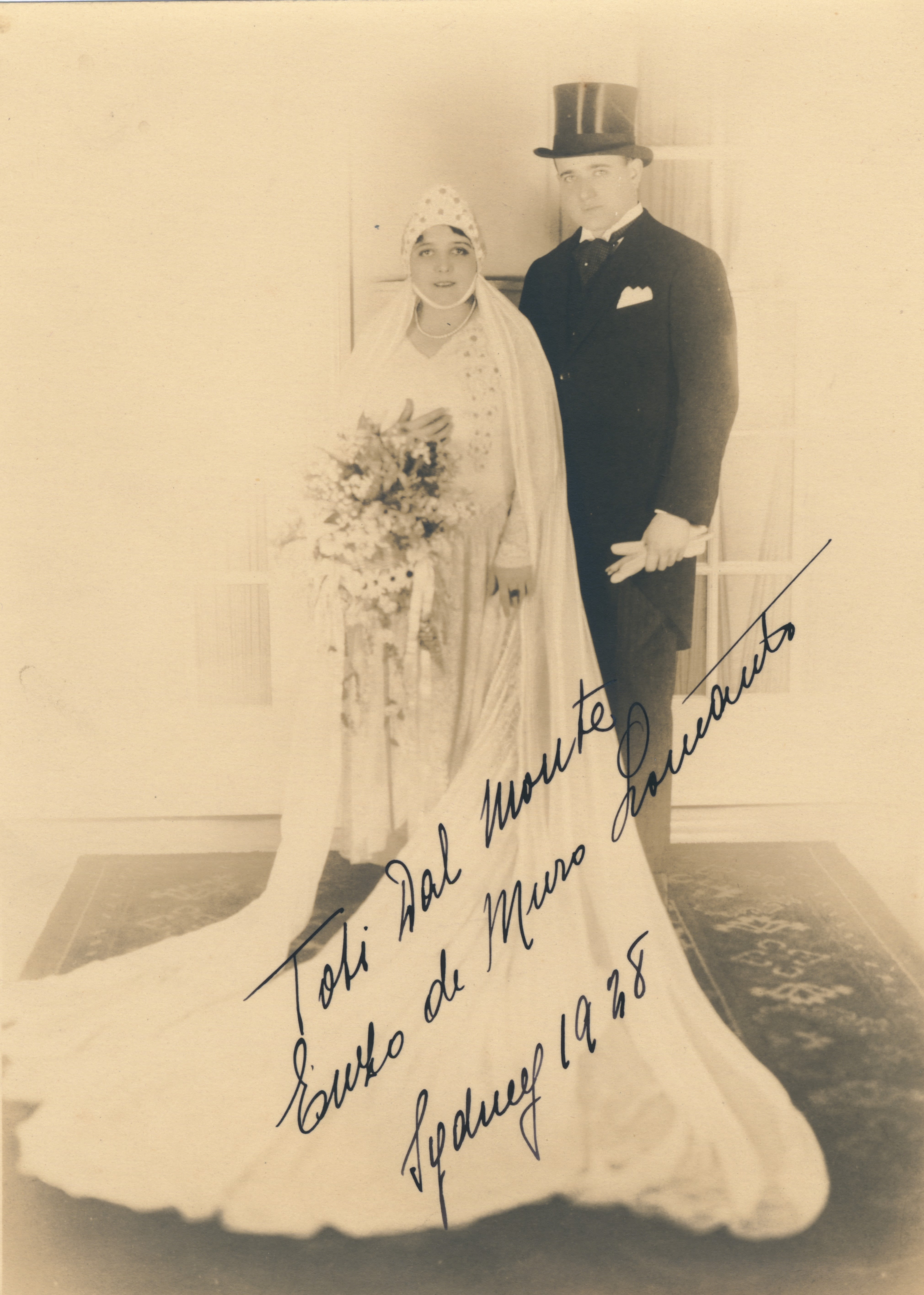 Full length portrait of a married couple. Woman on the left is in a white bridal gown just above the ankle. The dress is long-sleeve and features embroidery on the bodice. She is wearing a bonnet headdress and veil draped around her shoulders that spills out on the floor in front of her. She is holding a large bouquet of flowers. The man on her right is wearing a suit and top hat. He is holding white gloves in his right hand. They are both looking at the camera and are standing on a persian-style rig with floral and geometric patterns. 'Toti Dal Monte Enzo de Muro Lomato Sydney 1928' signed using black pen over the front of the photograph. 