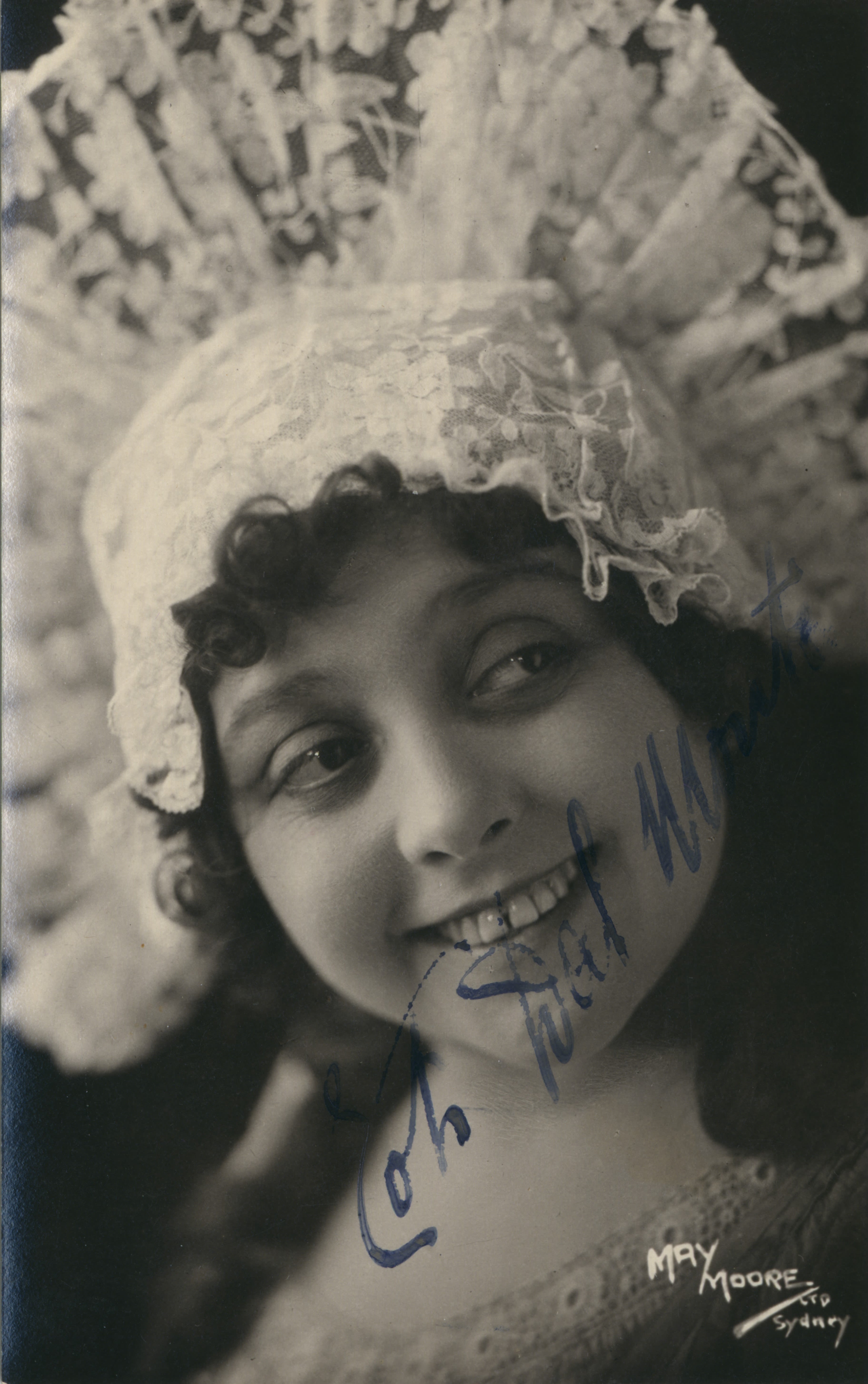 Head and shoulders portrait of a woman. She is wearing a tall headdress made of lace that fans out above her head on either side of her ears. She is facing towards the camera with her head tilted to the left and is looking towards the right. She is smiling. Hand written signature covers some of her chin and bottom-half of her face in blue cursive writing. 