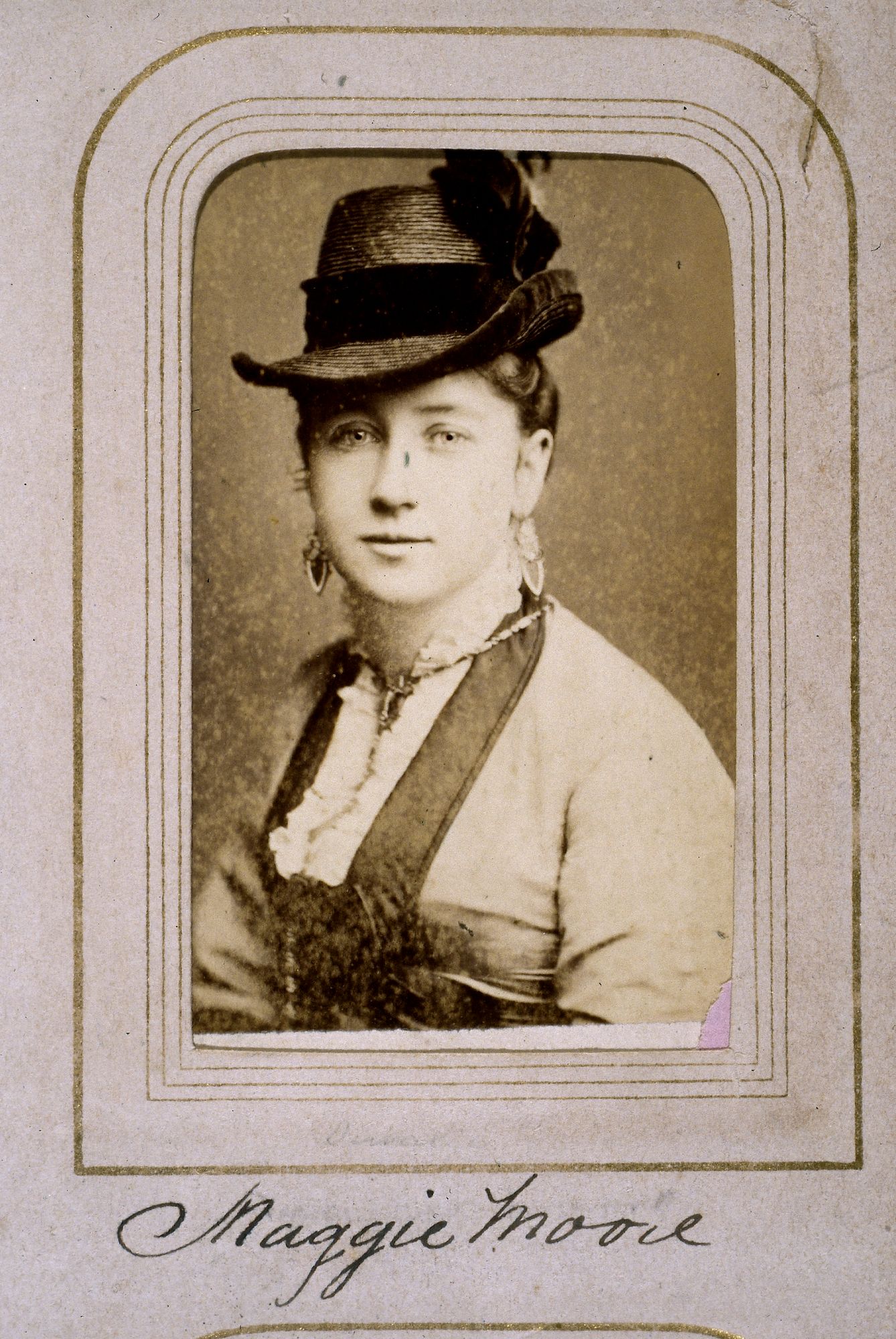 Head and shoulder portrait of a woman wearing a hat, dangling earrings and jewellery. Photo stuck inside scrapbook.  