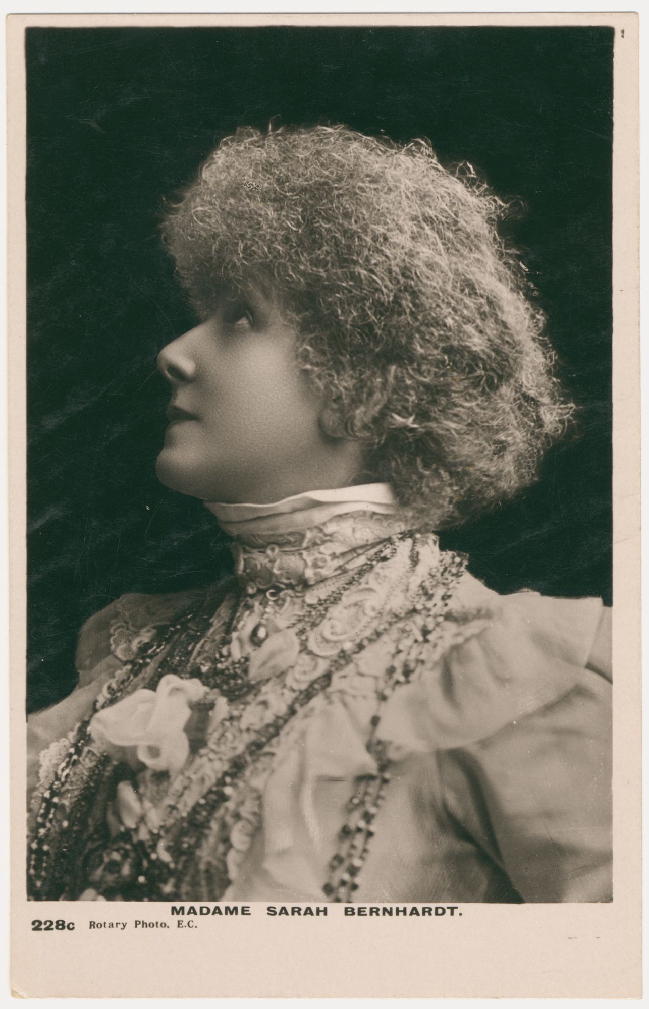 Head and shoulders portrait of a woman. She is wearing a nineteenth century style costume. The costume is made from white fabric including lace and is high-neck. Multiple beaded necklaces drape her neck. She is turned to the left with the profile of her face in view, and she is looking up to the top left corner of the image. She has curly hair and is framed on a black background. 