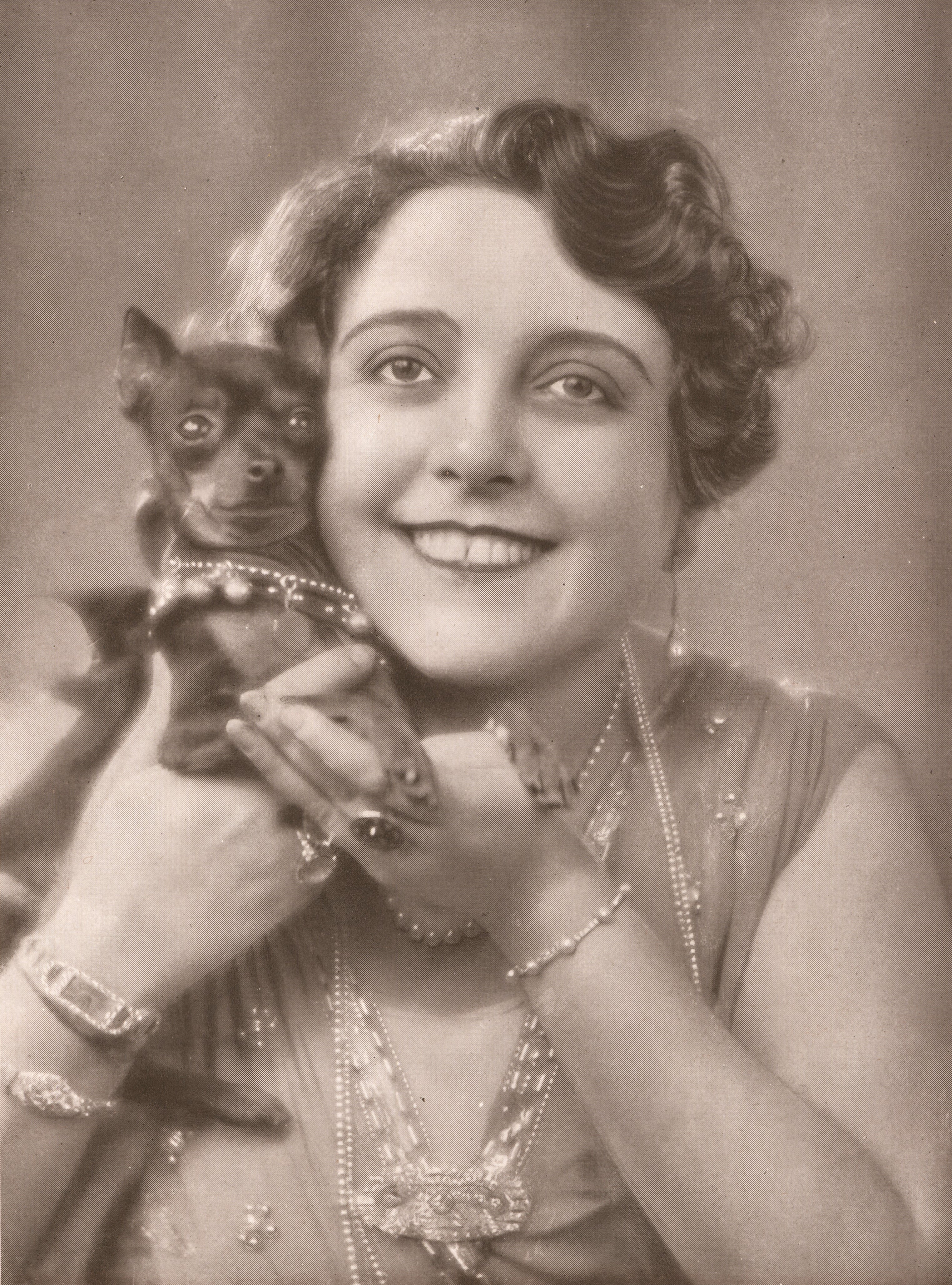 Portrait photograph of woman. She is wearing a sleeveless dress embroidered with jewel and sequinned embellishments and is wearing pearl necklaces, a pearl bracelet, and diamond watch. She is smiling and looking just above the camera. Her hair is pinned back and she is wearing dangling earrings. To her left she is holding a small dog. The dog is wearing a pearl collar and is looking to the right of the camera. 