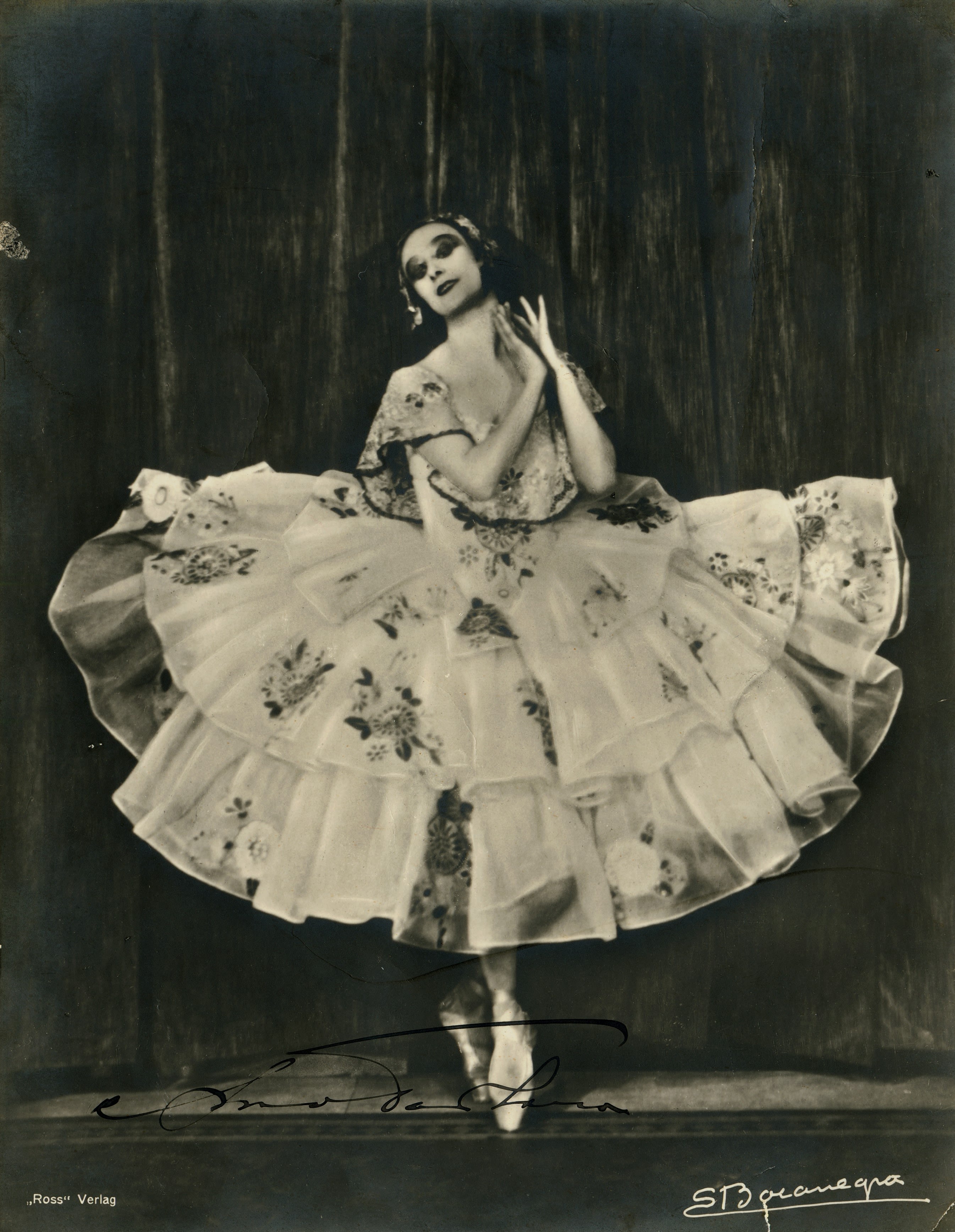 Full body portrait of a woman wearing a tutu dancing on stage. Her arms up raised and her hands are almost touching next to her chin on the right. She is in front of a theatre curtain.  The tutu is ankle length and is spread out to almost the width of the frame. The costume is light coloured with flowers embroidered all over the costume. She is wearing ballet shoes and is en pointe.  