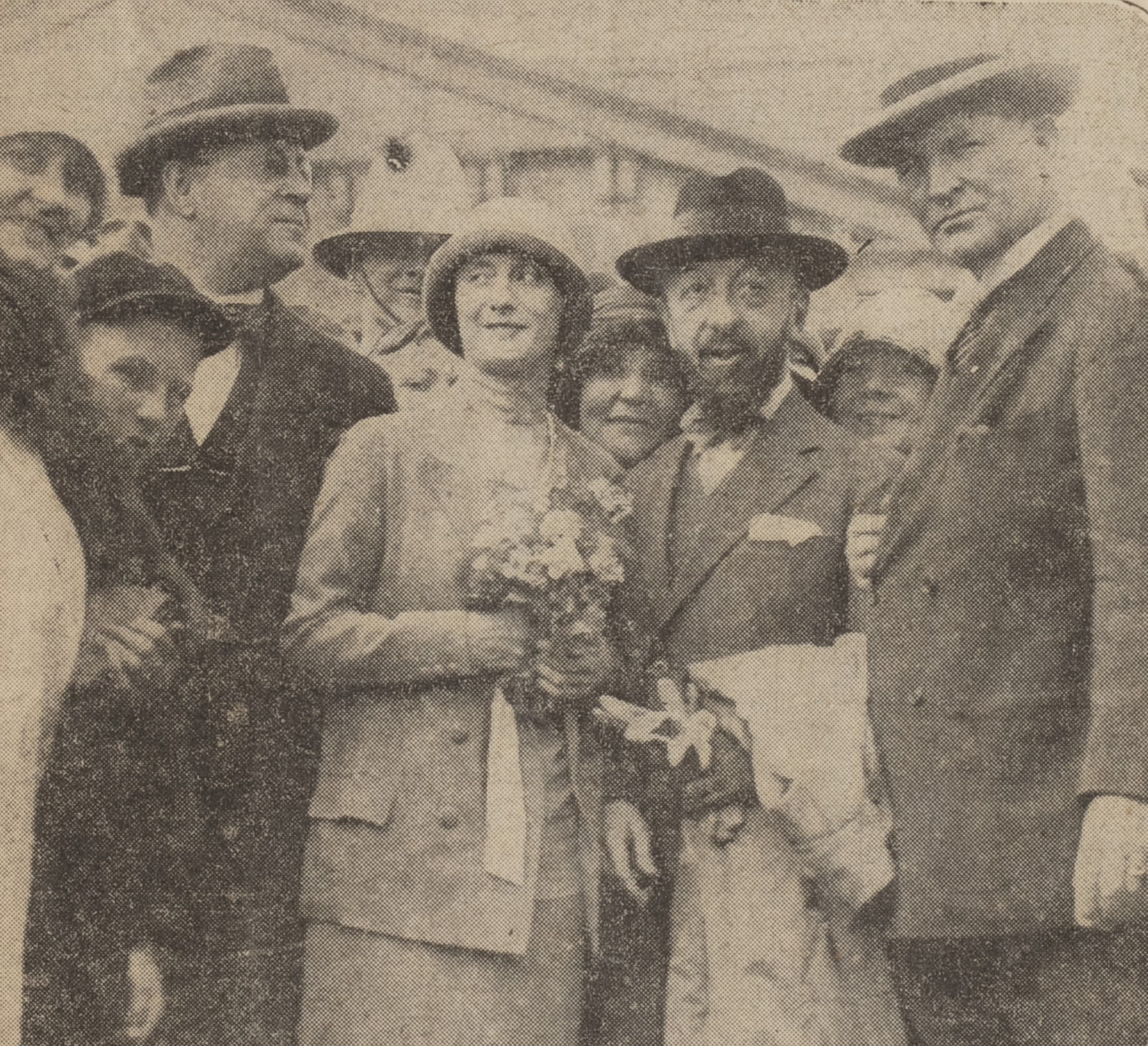 Photograph of nine people crowded around a woman holding a bouquet of flowers. The men a wearing suits and top hats, and the woman holding the flowers is wearing a light-coloured suit with a long skirt and bowel hat. Some people are looking towards the camera. Photograph in newspaper and has grainy appearance. 