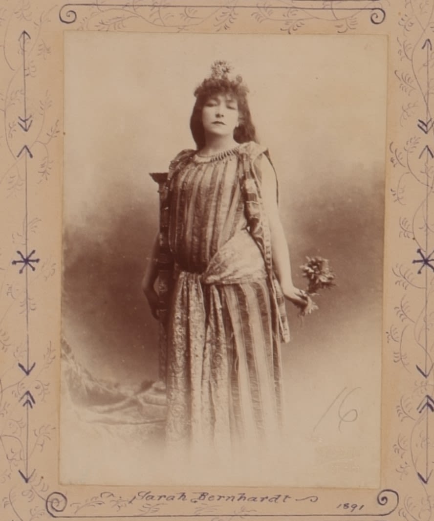 Portrait of a woman from her head to just amove her ankle. She is wearing a theatre costume. The costume is floor-length with vertical stripes and a thick fabric belt that is tied around her waist. She is holding a small bouquet of flowers in her right hand and is looking pensive at the camera. She has curly hair and is wearing a small headdress. Portrait is stuck in a scrapbook that is decorated with hand-drawn foliage in blue pen. 