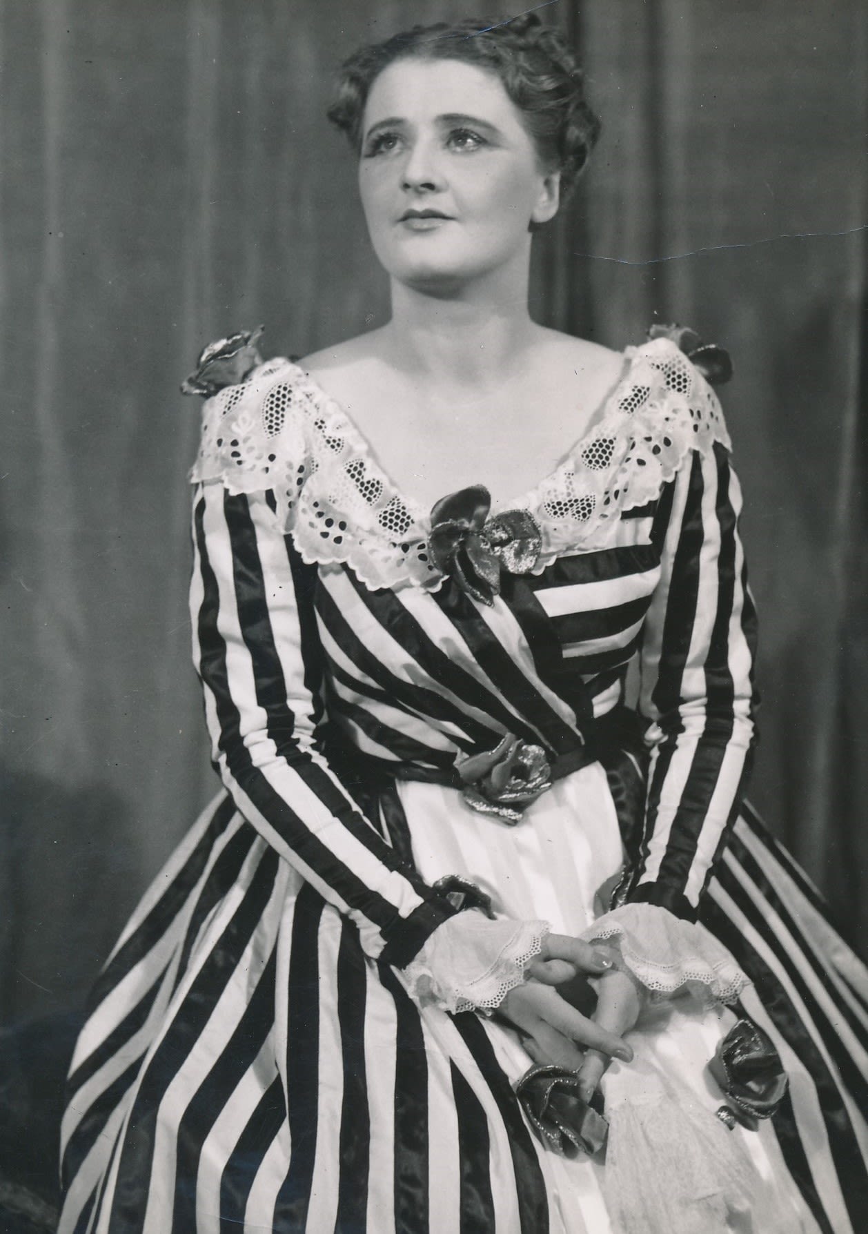 Photo of woman dressed in a striped costume with artificial roses in the centre of her neckline, waist, and shoulders. She is looking up towards the left corner. 