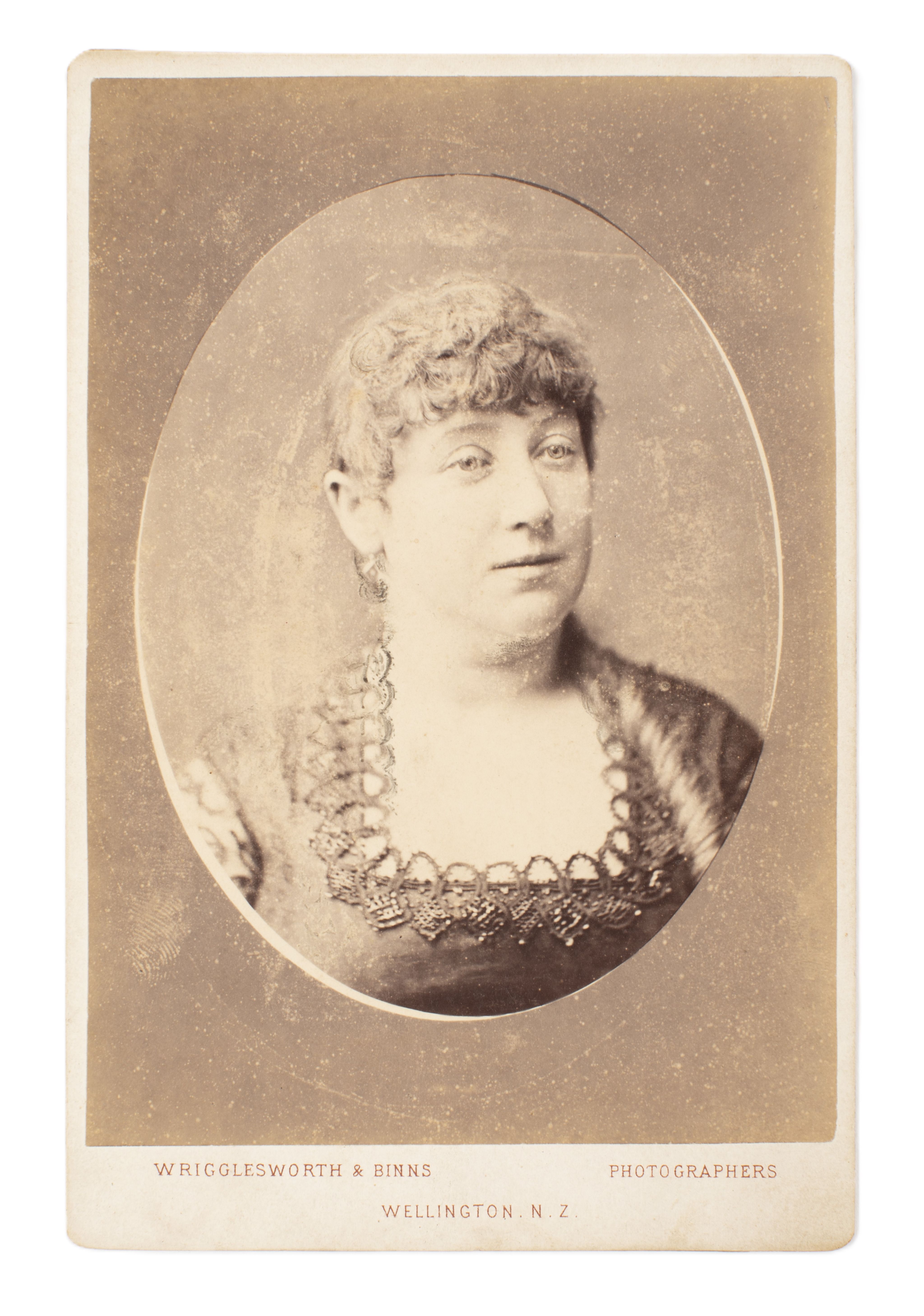 Head and shoulders portrait of a woman wearing a dress with a square neckline. Photo is framed in an oval window on a cabinet card. 