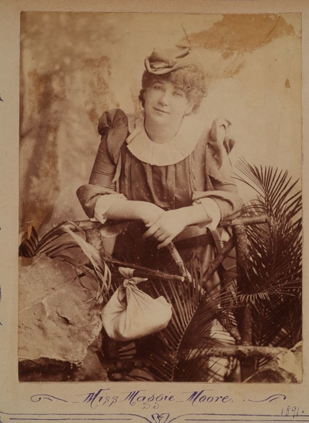 Portrait of woman from head to bottom of her knees. She is wearing a dress with puffy shoulders and white-frilled neckline and cuffs, and a small hat on the left side of her head. She is smiling without teeth at the camera. The woman is surrounded by ferns, rocks, branches, and is resting her forearms on a small fence-like structure. There is a piece of cloth wrapped up like a rucksack handing from the fence. Photo is stuck in scrapbook. 'Miss Maggie Moore' is handwritten in cursive writing down the bottom of the image. 