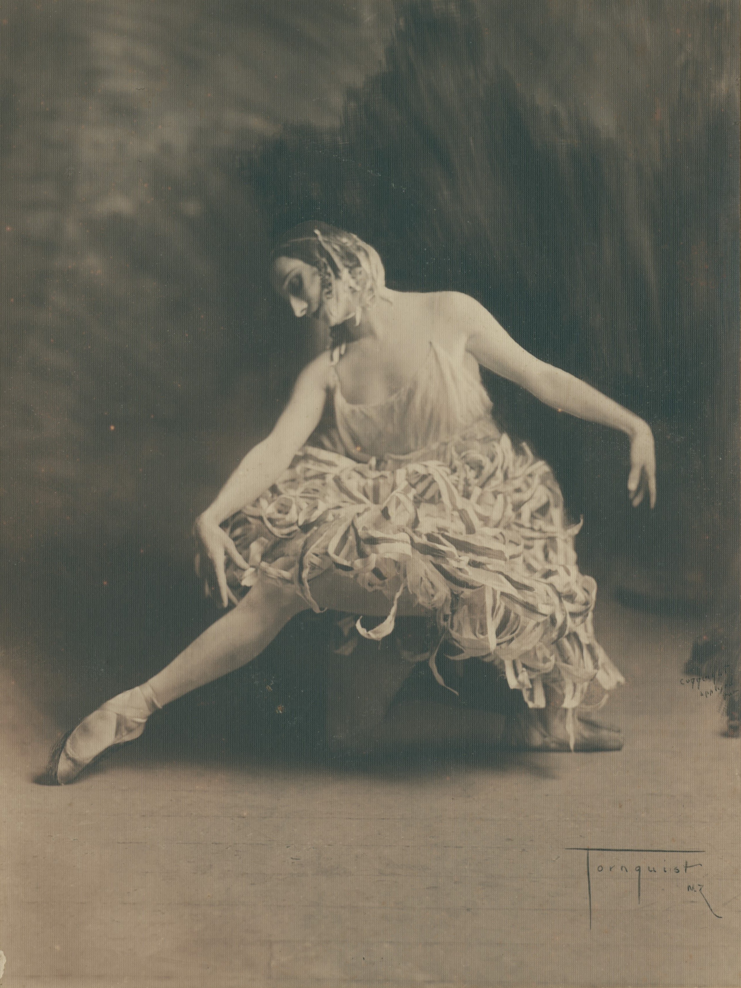 Full length photograph of woman. She is a dancer dressed in tutu that is made from strips of fabric. She is kneeling with her right leg crossing her body to the left, with her right foot en point. Her arms are draped to either side, and her head is tilted to the left and she is looking towards her right foot. Inscribed with the photographer's signature on the bottom-left of the image. 