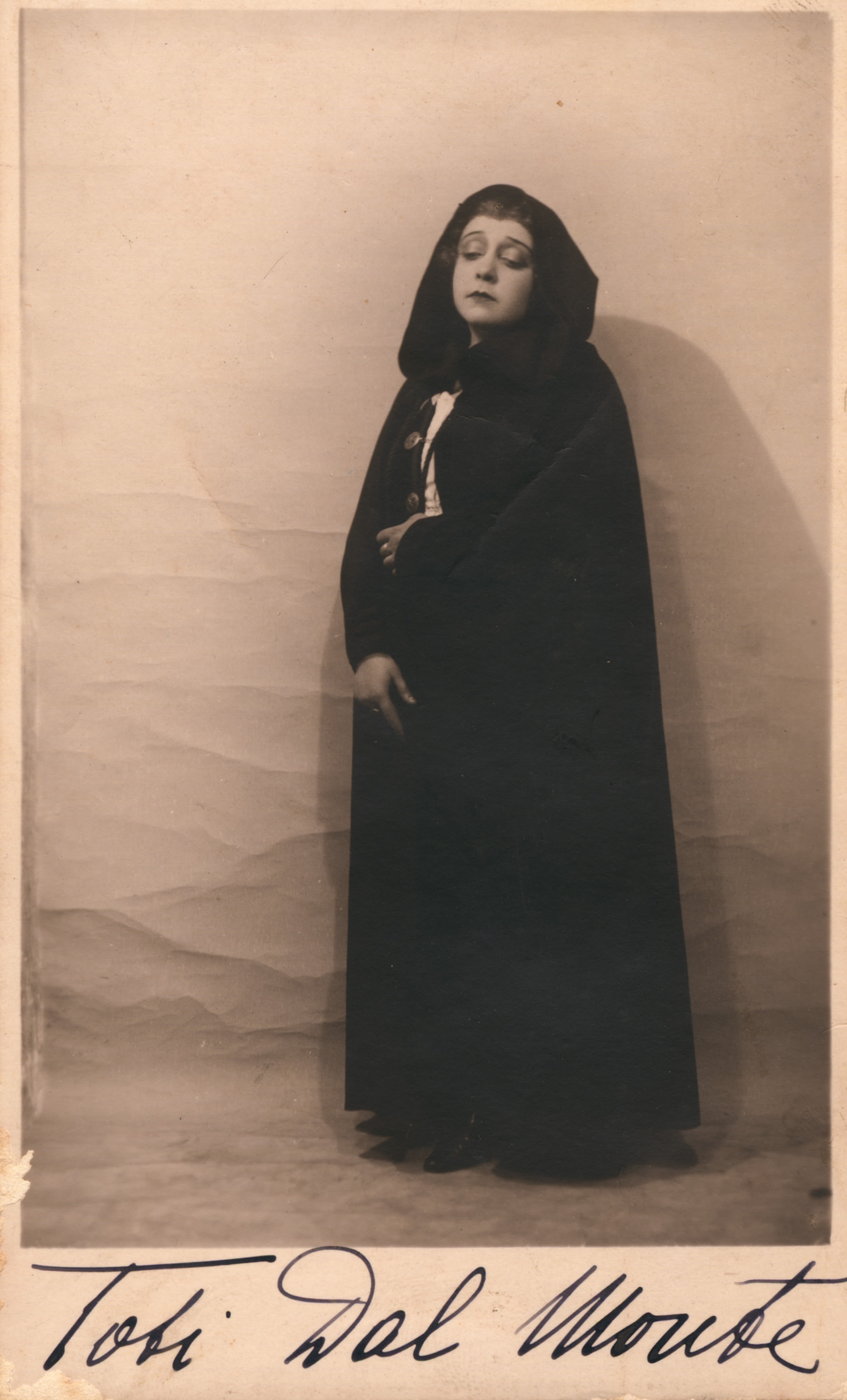 Full length portrait of a woman. She is wearing a floor-length hooded robe and is slightly right of frame. Her eyes are closed and she has a sombre expression. Underneath the image is 'Toti Dal Monte' written in black cursive writing. 