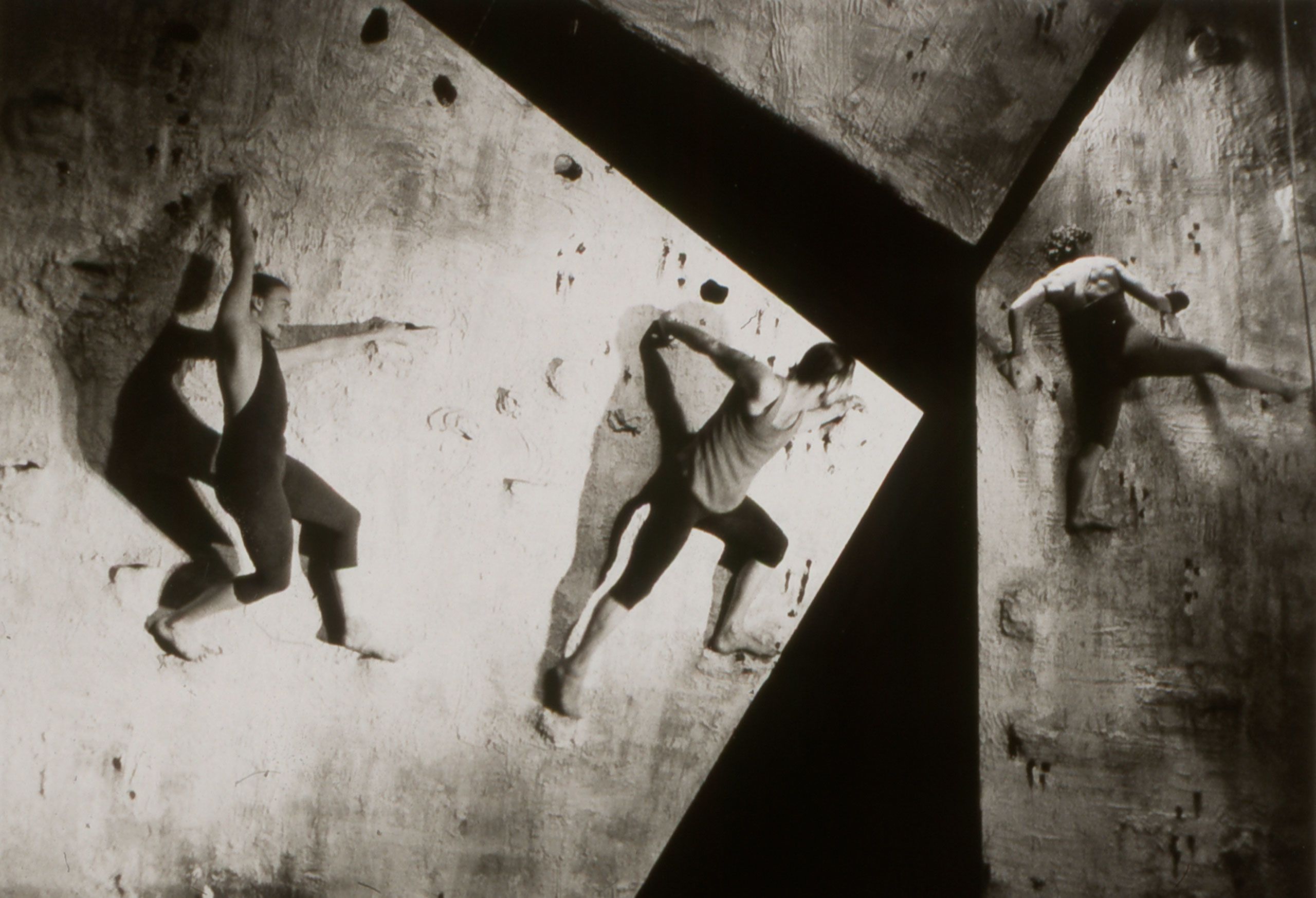 Three performers wearing leggings climbing across two angled walls. Black and white photo shows strong contrast and angular shapes.
