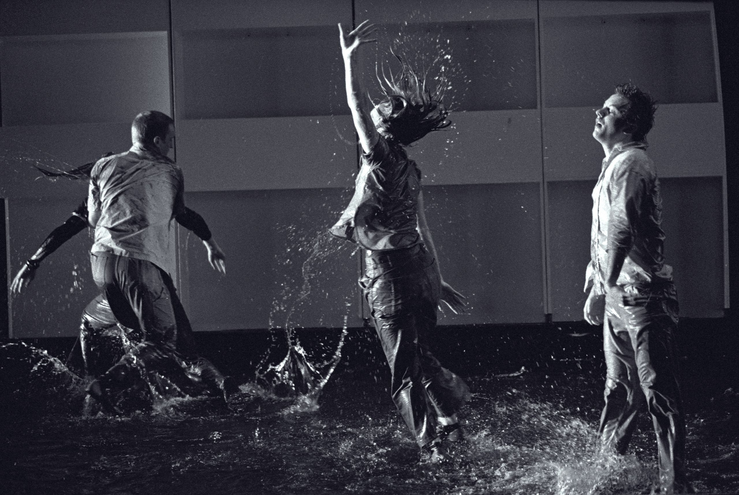 Four dancers perform on a stage covered in water, making splashes and droplets fly up.