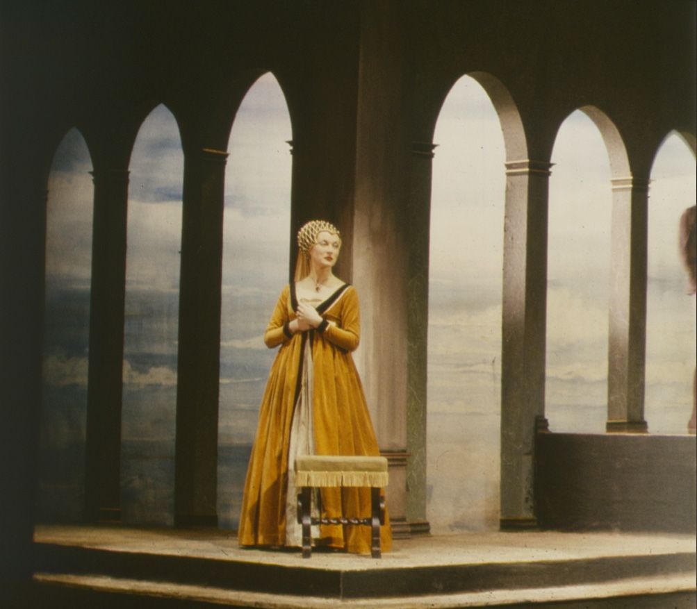 A solitary female actor wearing middle ages style costume, stands with hands clasped at her chest, looking into the distance. A backdrop of archways shows a landscape in the distance.