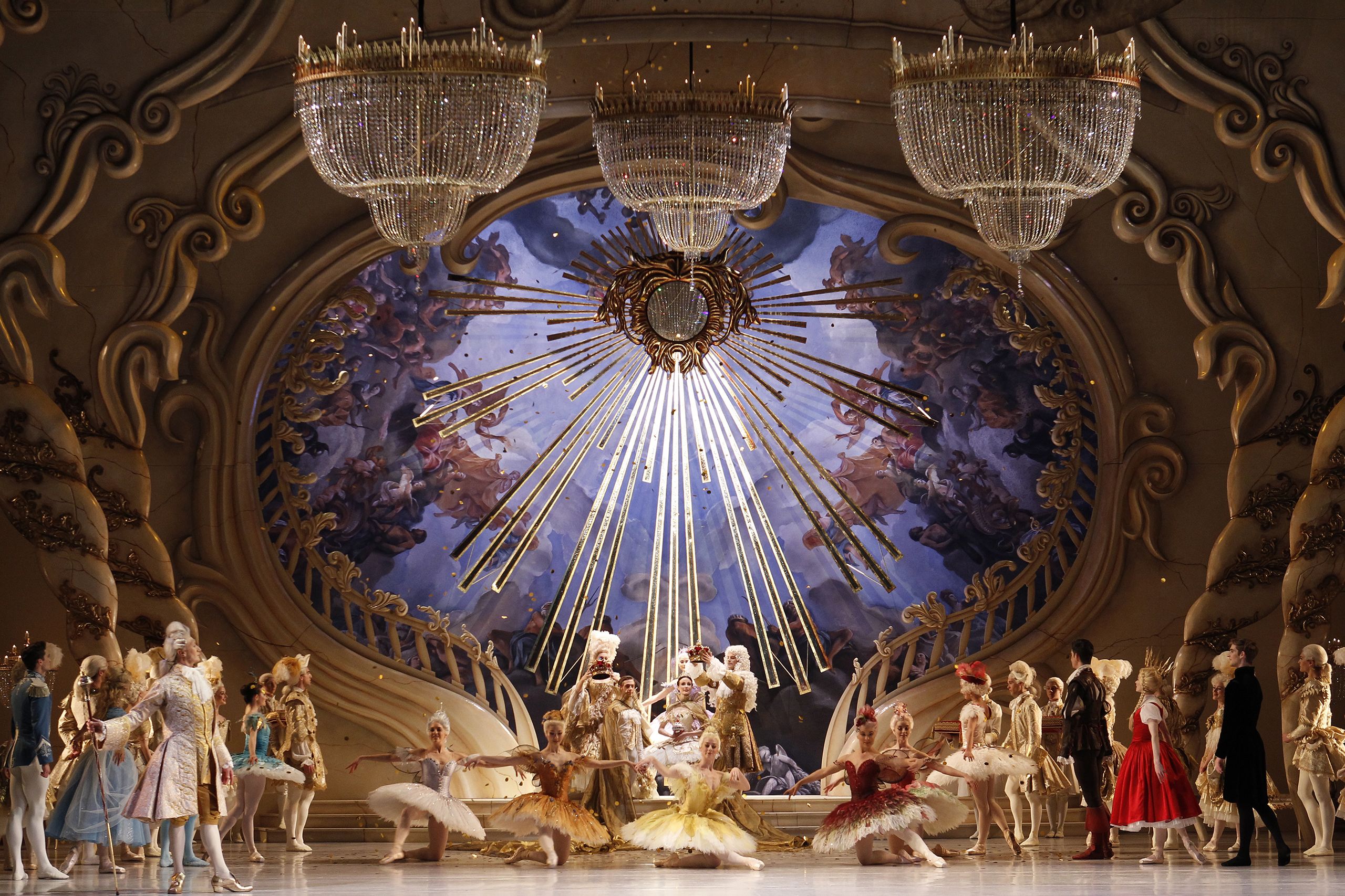 An elaborate Baroque style set featuring a sweeping circular staircase balcony and large starburst chandelier above the ballet dancers on stage. A circle of dancers in the centre with members of the corps de ballet looking on from each side.