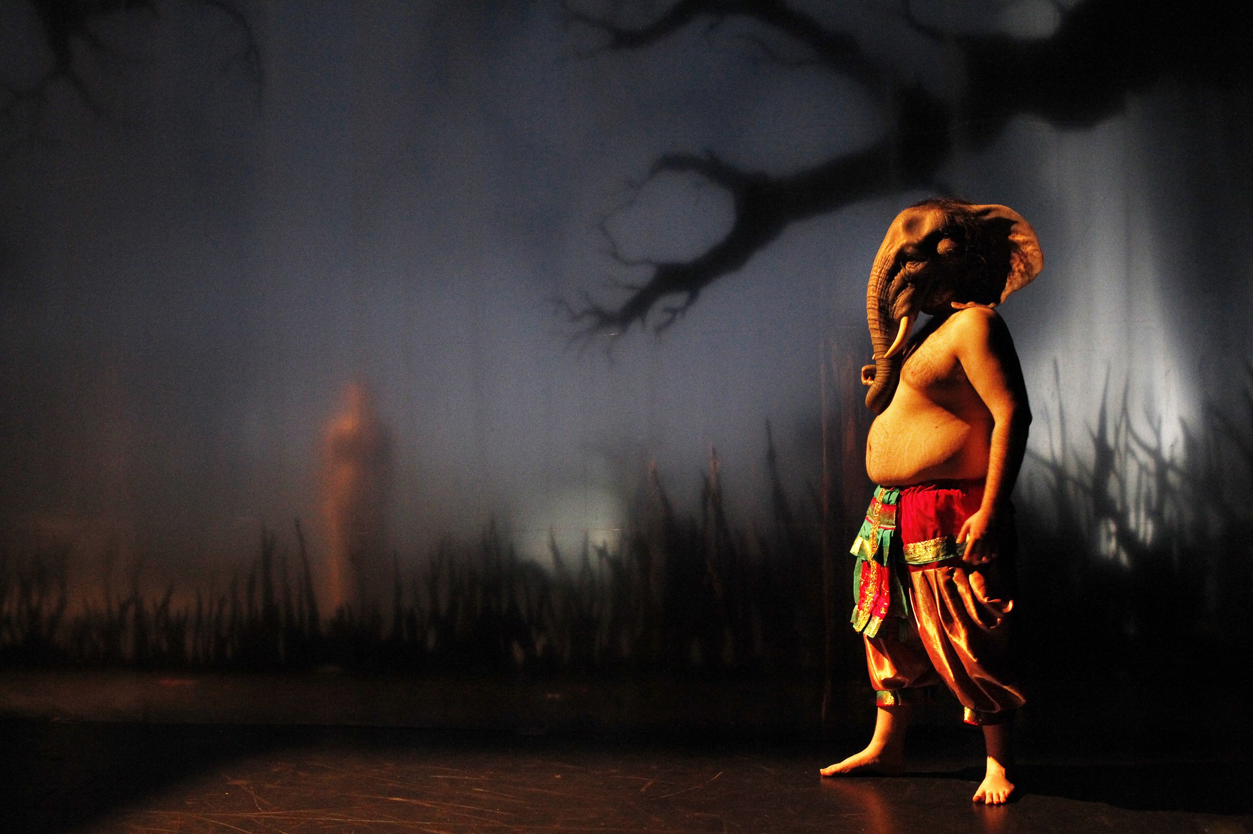 Actor wearing an elephant head costume, bathed in yellow light. The set appears foggy with gnarled tree branches and long grass.