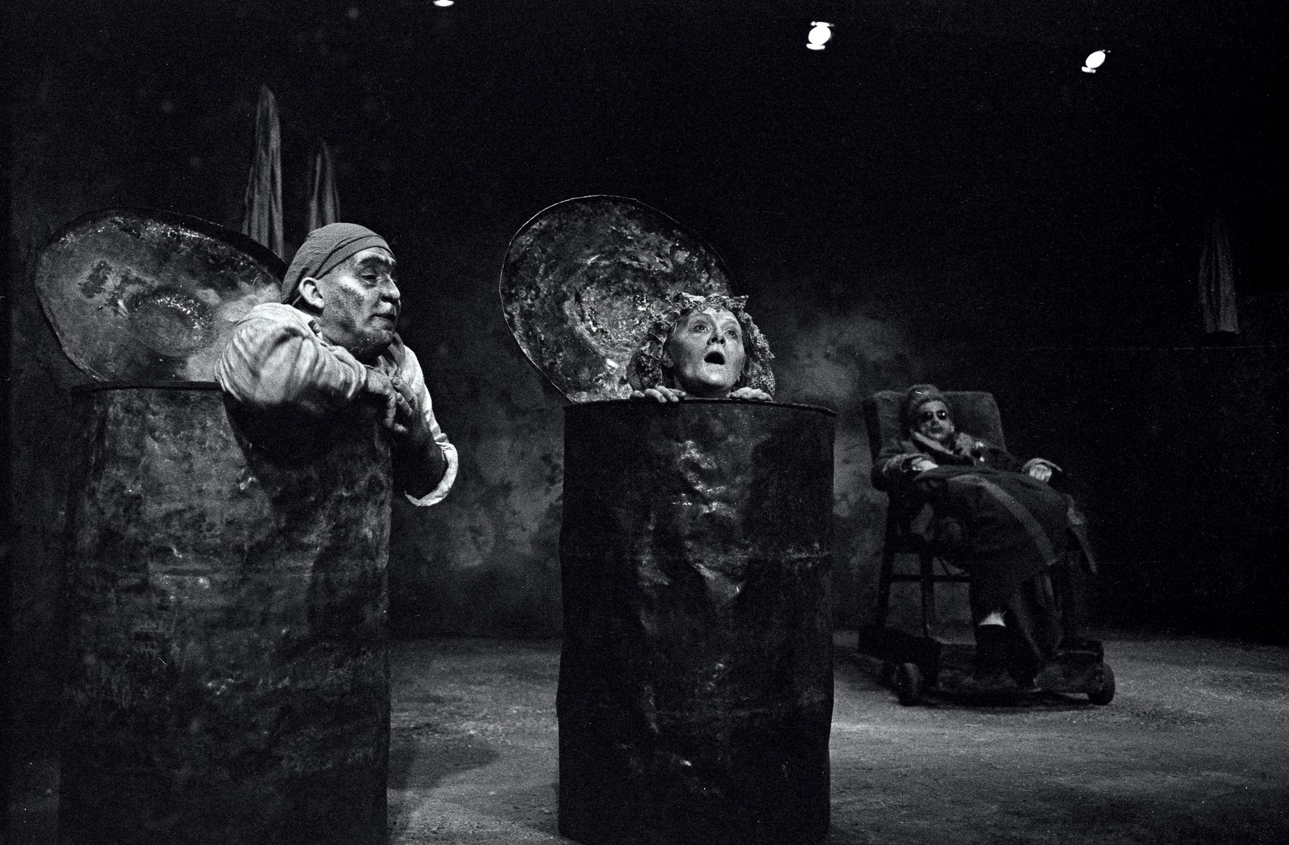 Black and white photograph of two actors, each emerging from a 44 gallon drum looking disheveled. A third character sits in a wheeled chair in the background. The set is gloomy.
