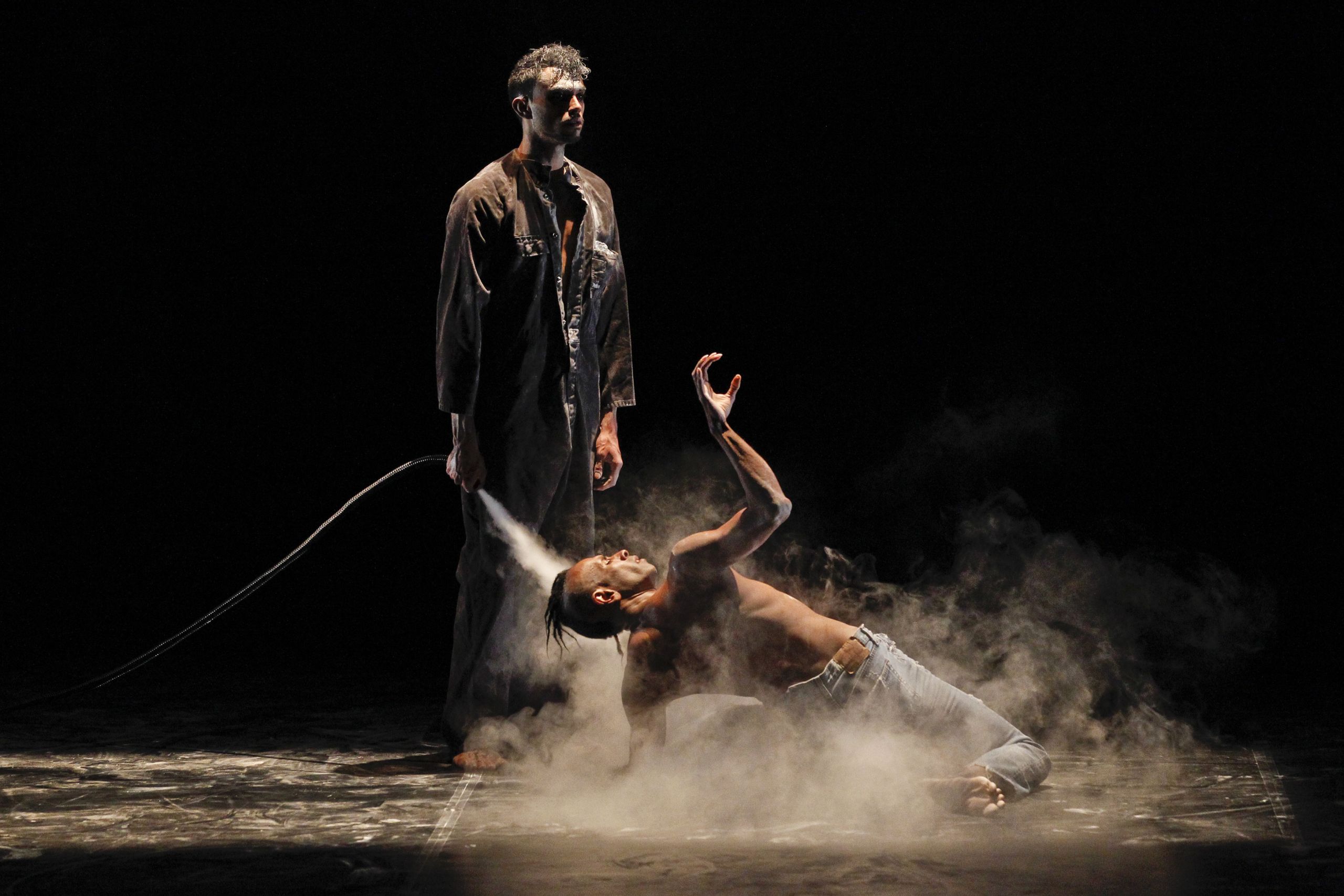 A male dancer twists on the floor while another man stands and directs a stream of smoke haze at him. Background is total darkness.