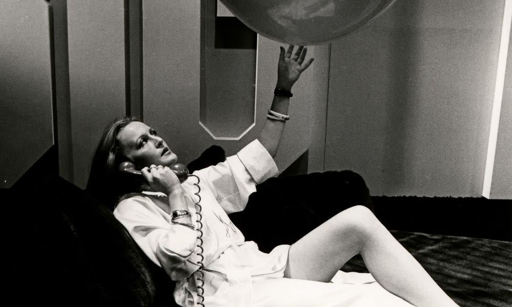 Female actor in a white robe reclines on a luxurious sofa holding a telephone receiver to her ear and a large balloon in the other hand. The phone itself is in the foreground.