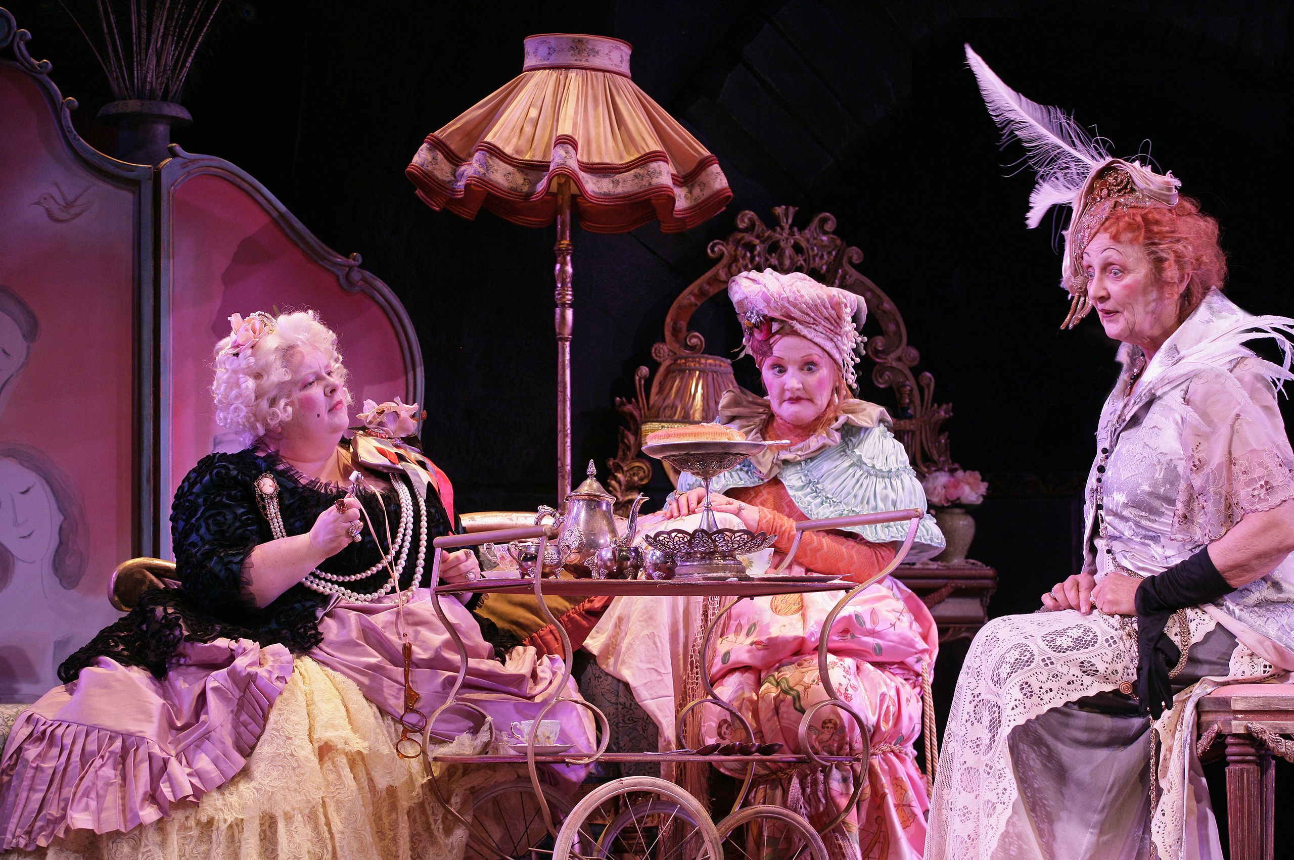 Three female actors in frilly, lacy dresses sit around a tea trolley with lively expressions.