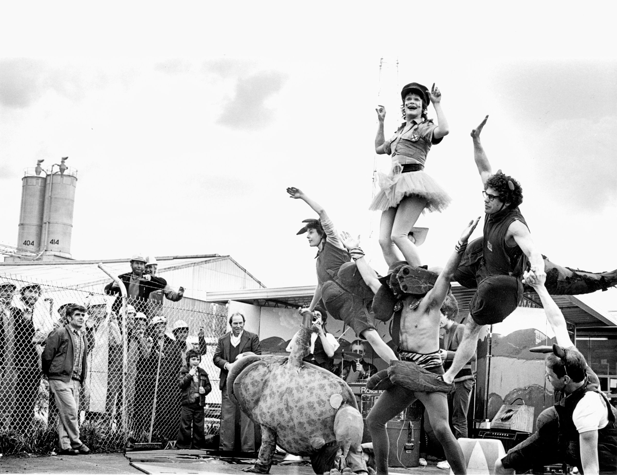 Six circus performers in humourous costumes balancing in a human pyramid outside near a factory. Workers in hard hats look on from behind a fence.