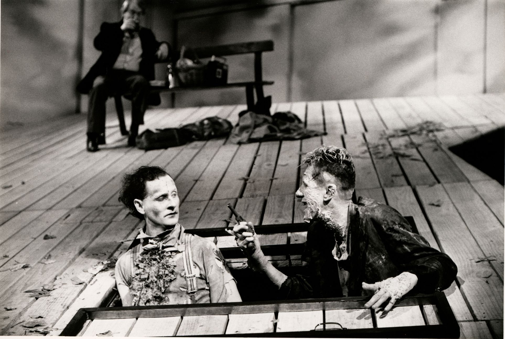 Two actors emerging from the floorboards, looking disheveled. One man holds a pistol. Black and while photo has a long persepctive behind the actors of the floorboards continuing into the distance.