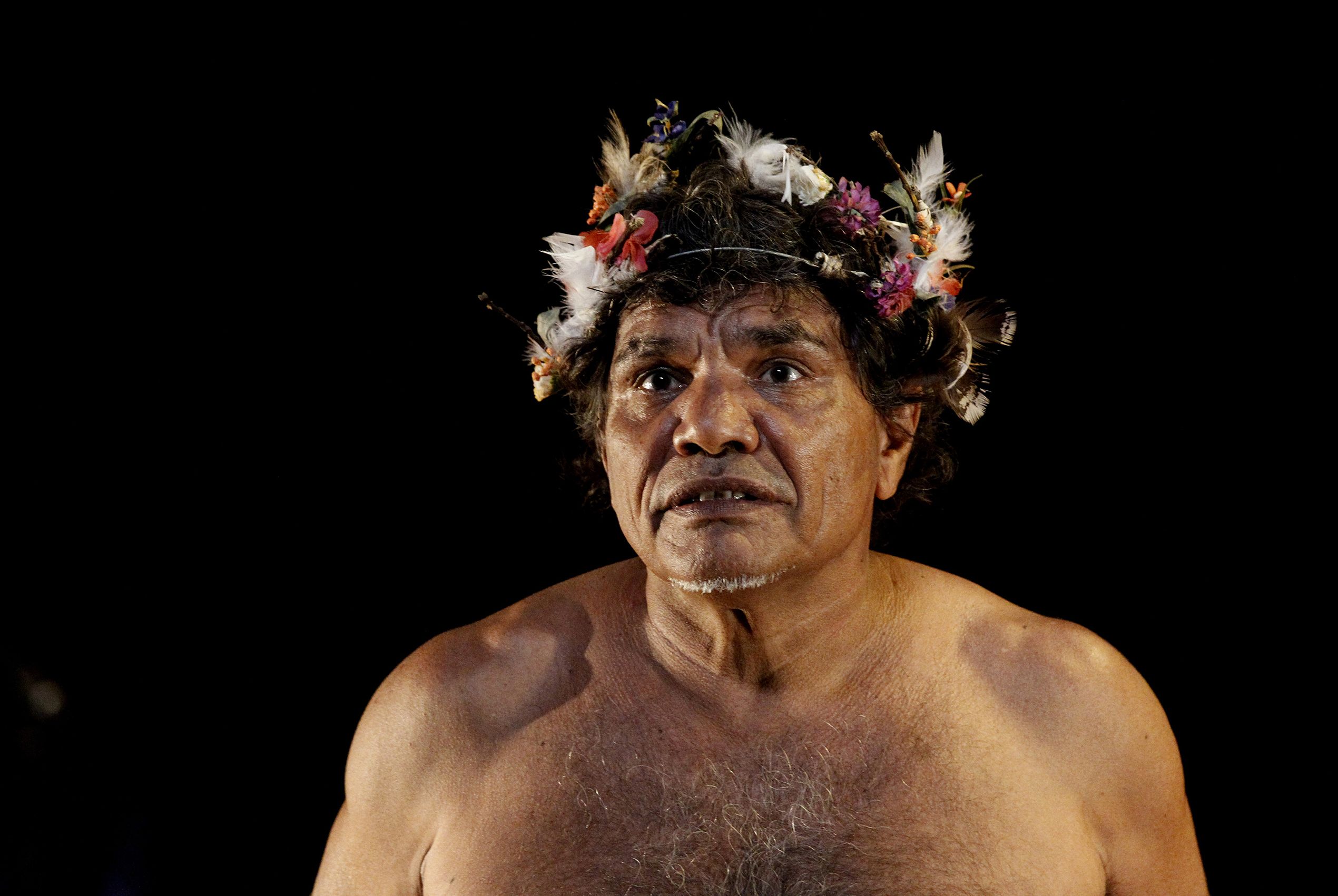 Close up of a shirtless indigenous actor wearing a small feathered headpiece. His expression shows concern.