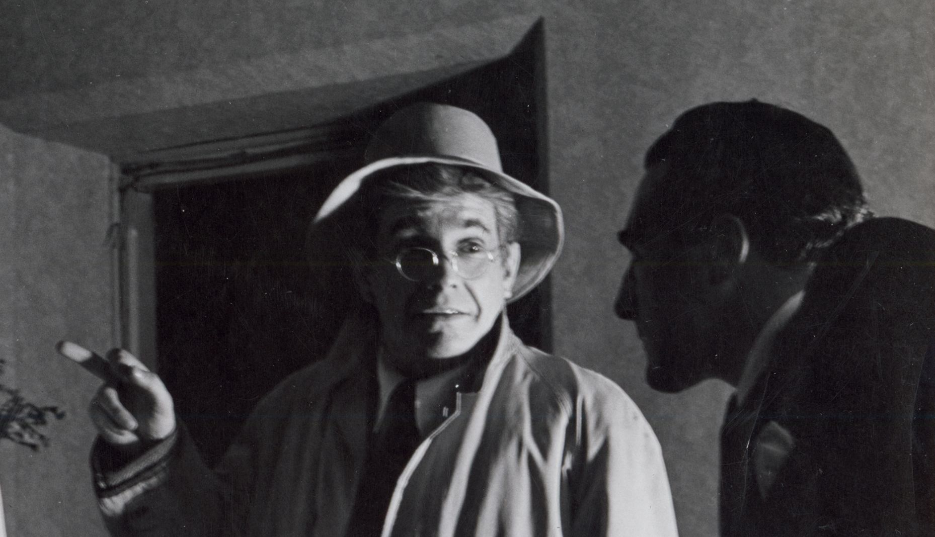 A male actor on the right appears as a dark sillhouette, looking at two other actors. The man in the centre looks surprised and points at a woman on the left. They are wearing 1940s era clothing.