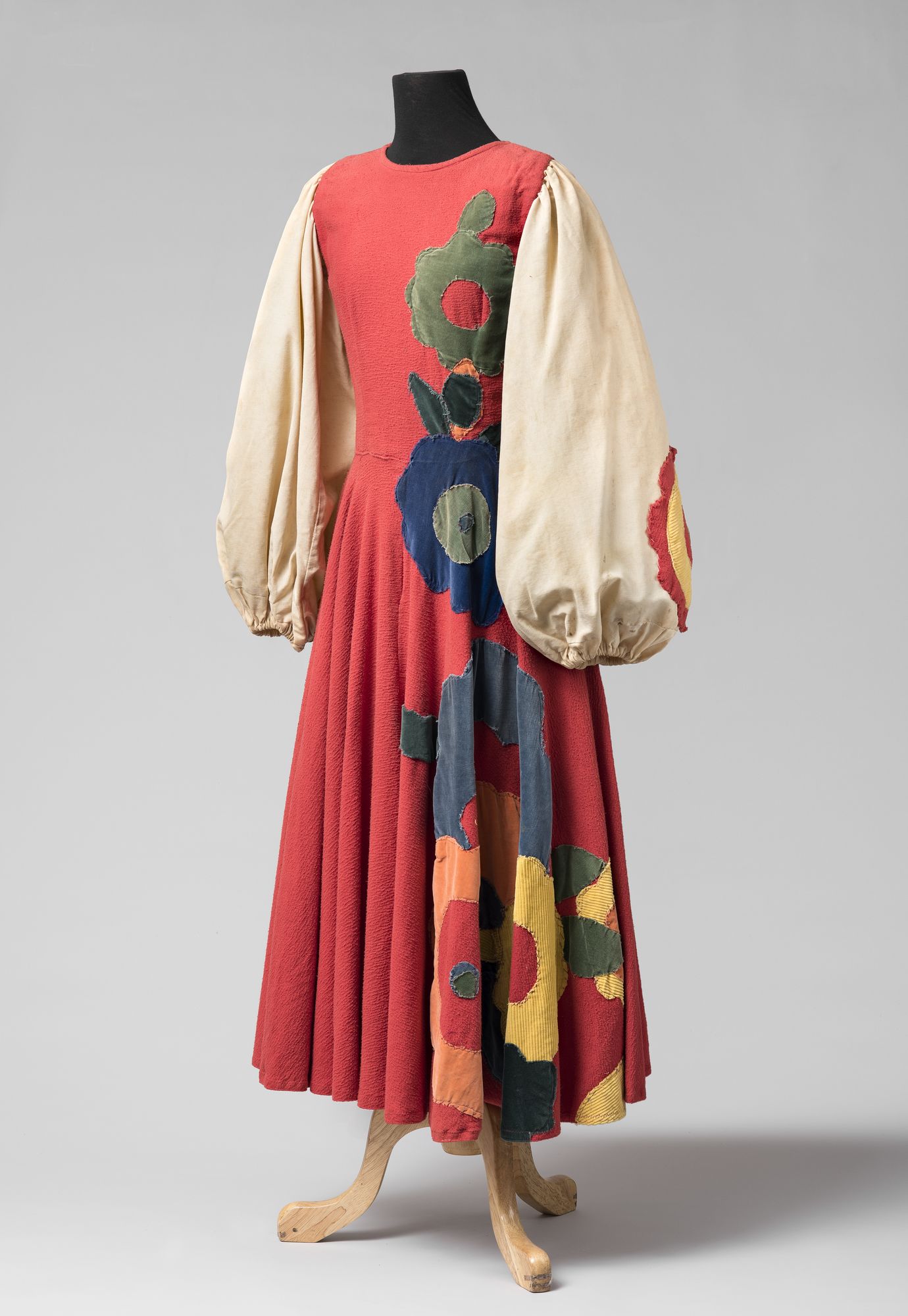 Colour image of a dress on a headless mannequin on a white background. Dress has large, cream-coloured balloon sleeves and a contrasting deep orange bodice and full skirt. Large floral applique is featured on the right side of the skirt and part of the sleeve.