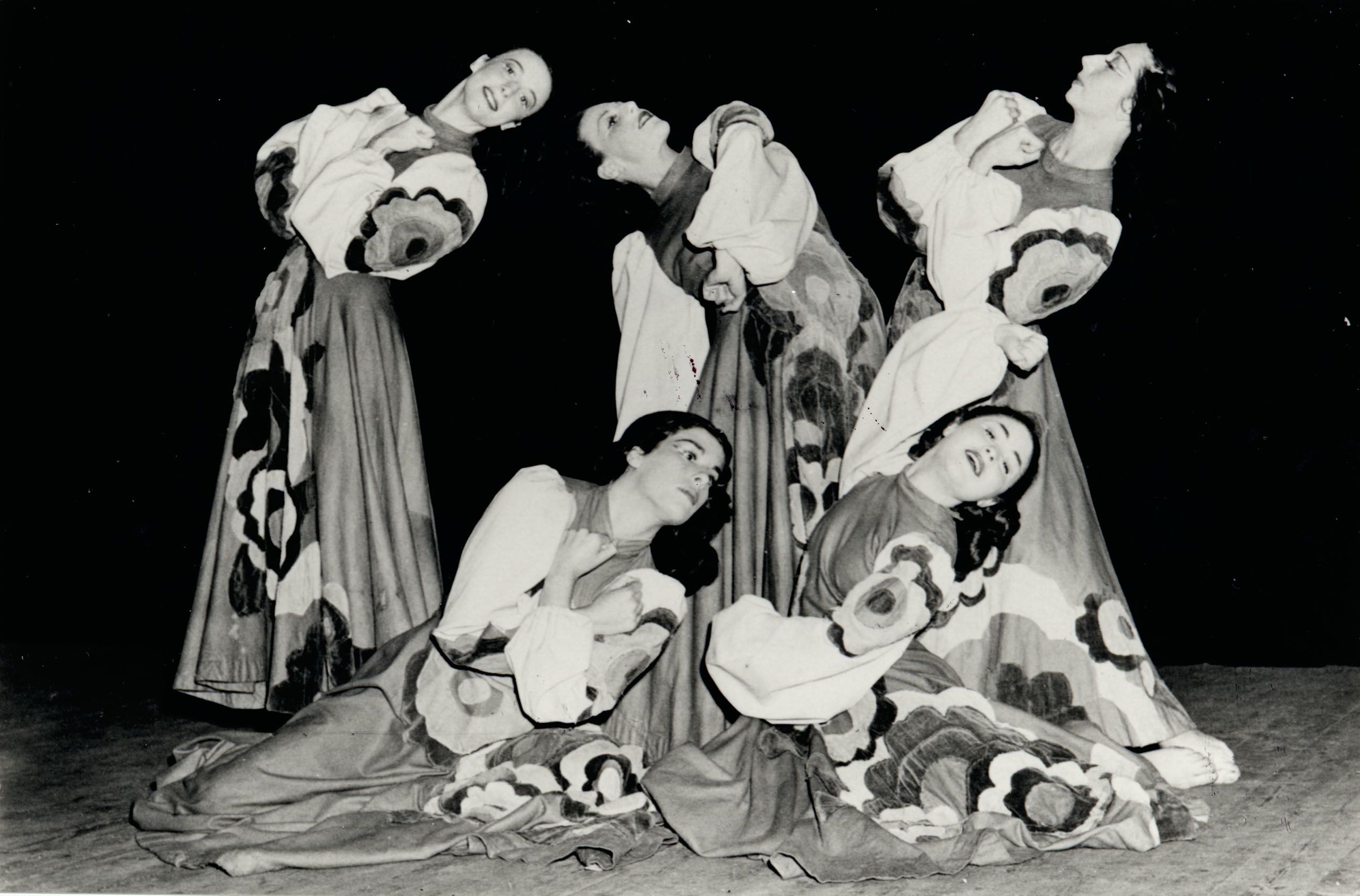 Black and white image of five female dancers wearing dresses with balloon sleeves and full skirts featuring large floral applique. Three dancers are posed standing leaning back with arms crossed to their chests, and two dancers in front are sitting on the ground with the same poses.