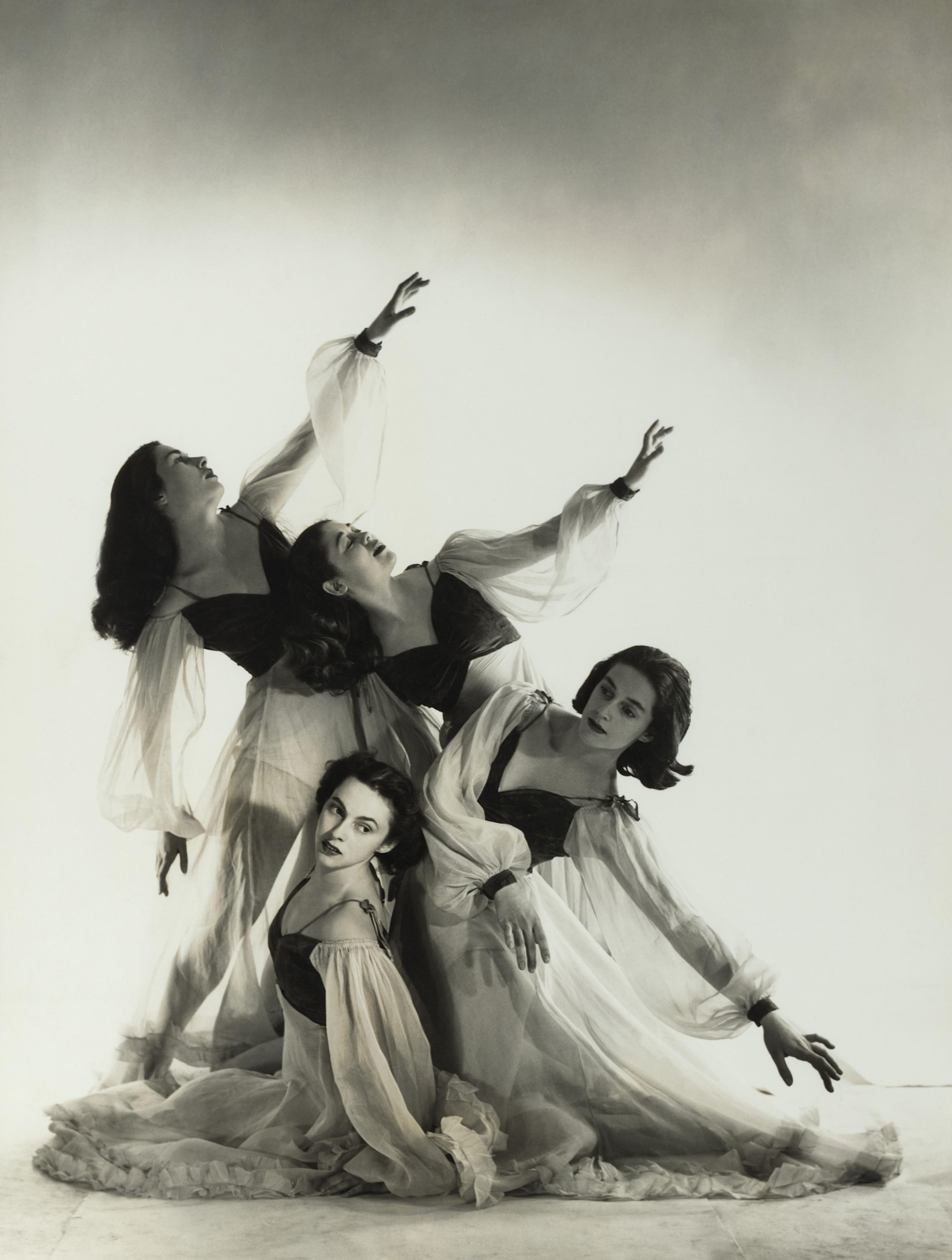 Black and white image of four dancers in standing and seated formation, with arms out-stretched upwards. Dancers are wearing flowing long sleeve gowns with black fitted bodices, on a white background. 